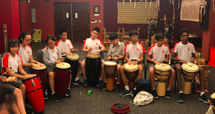 Eight students in white and red uniforms sit in a row playing djembe and conga drums in a red room.