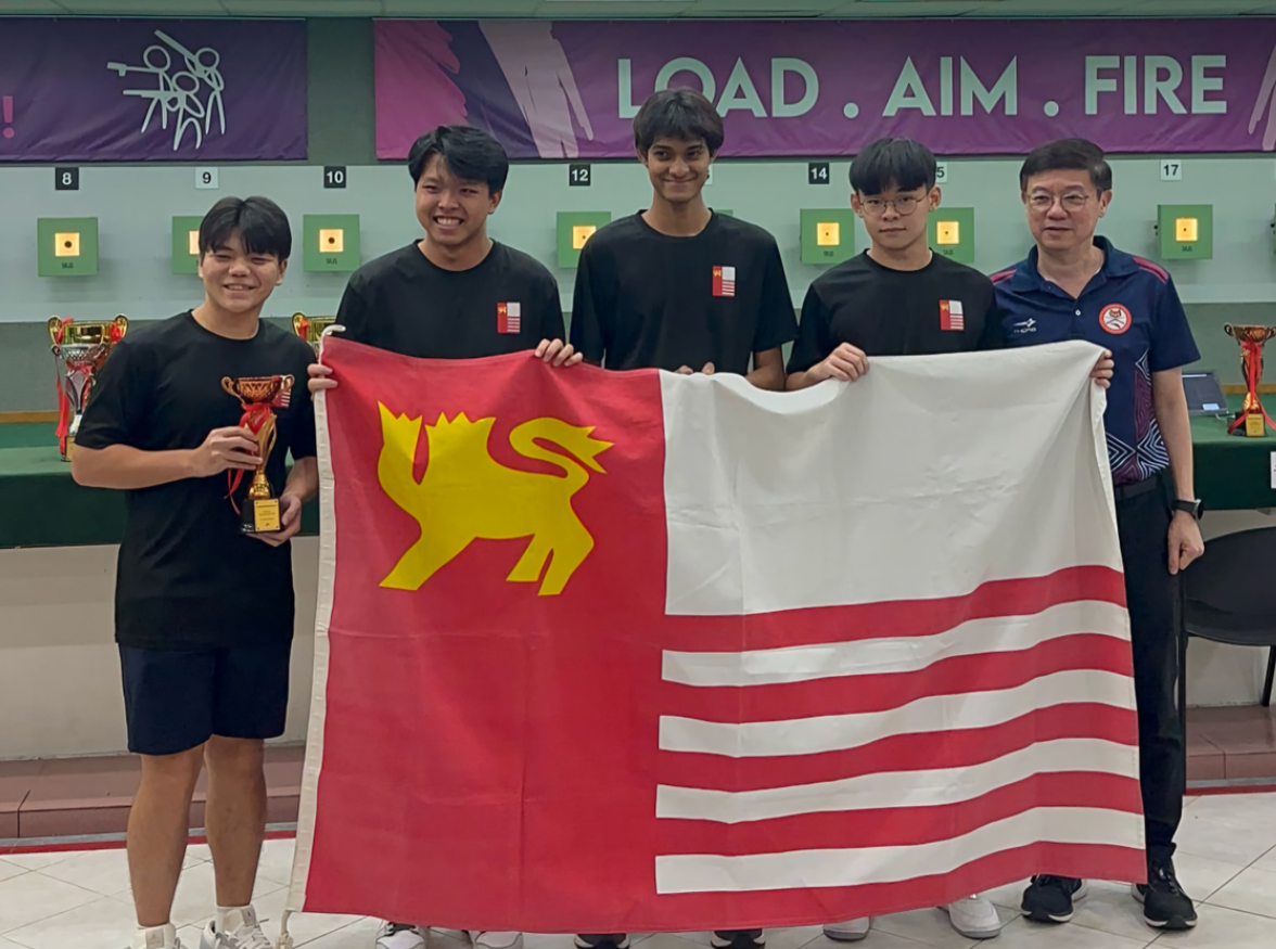 Five people stand at a shooting range holding a red and white flag with a yellow lion, under a "Load. Aim. Fire" banner.