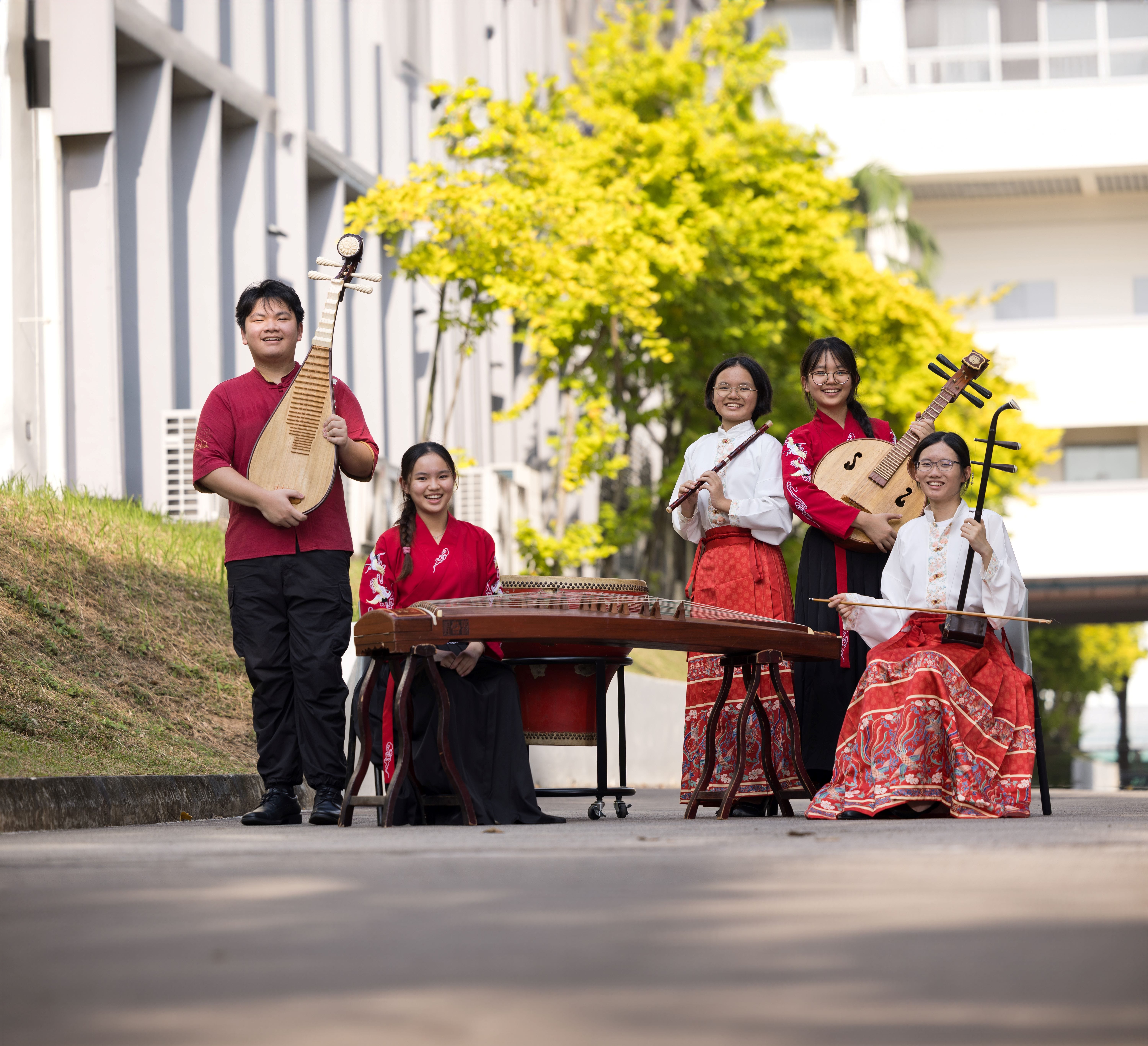 Five young musicians in red and white traditional Chinese attire pose outdoors with instruments including pipa, guzheng, and erhu.