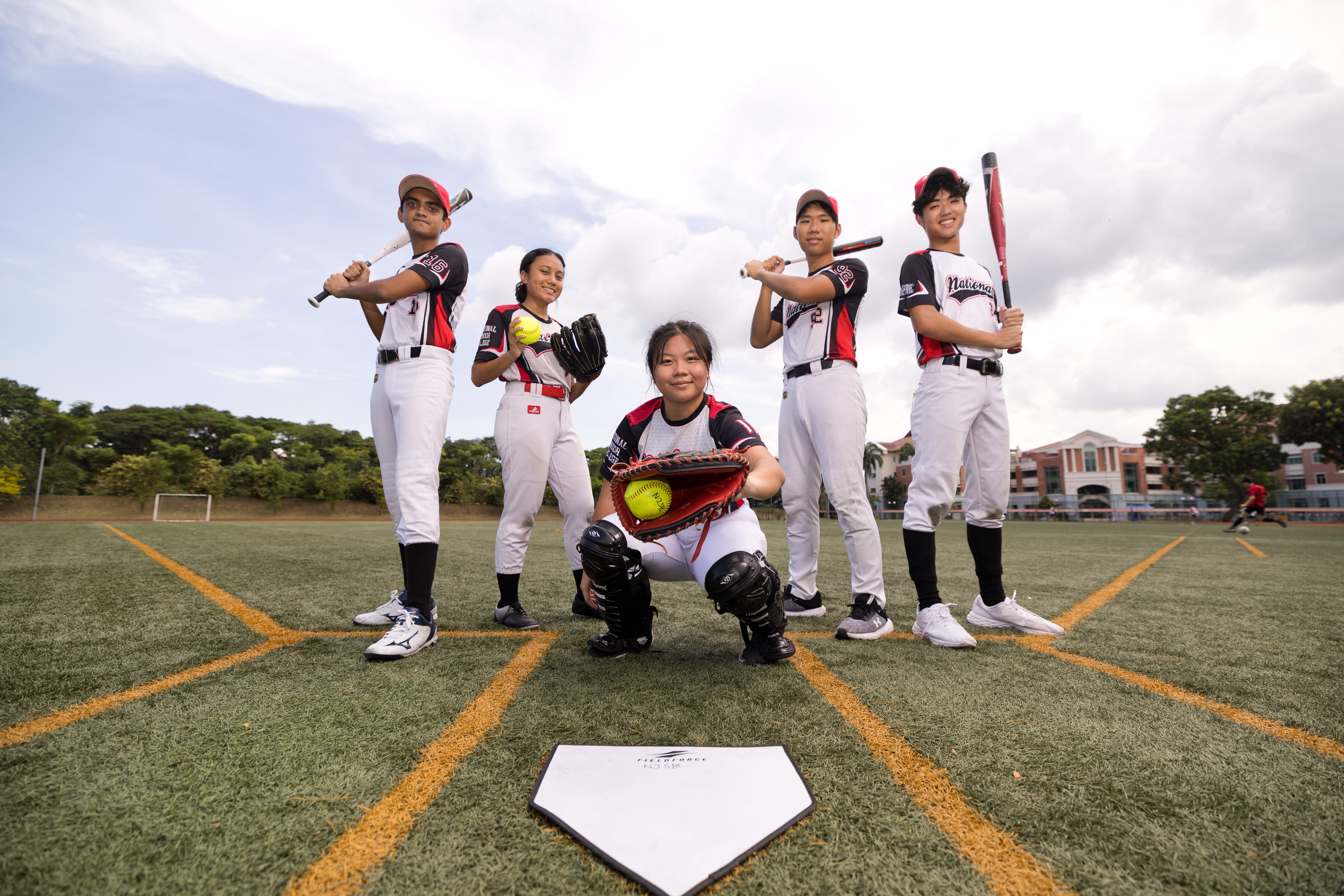 Five baseball players in uniforms pose on a field; catcher squats with glove holding a yellow ball.