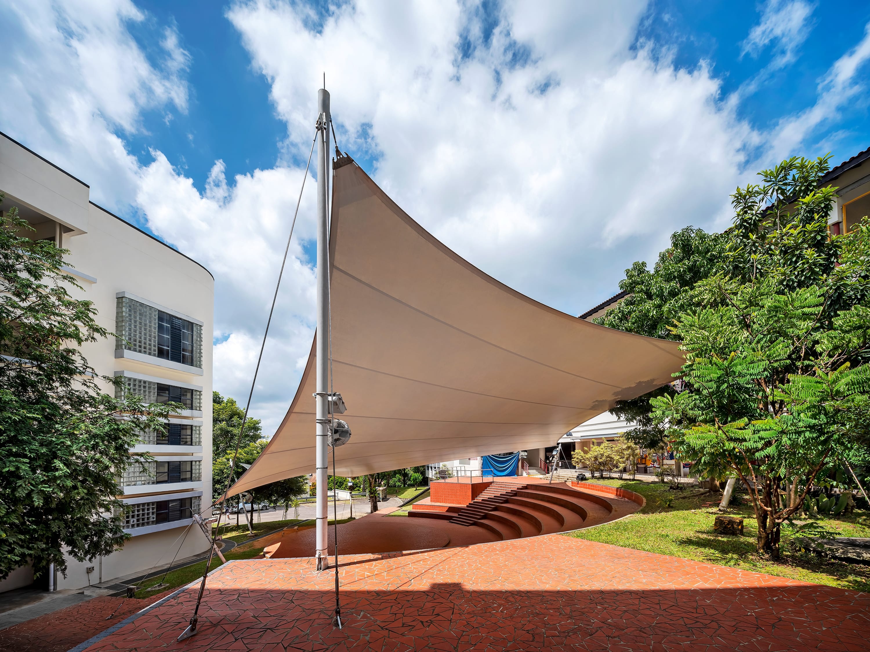 Large triangular shade sail over an outdoor amphitheater with curved steps, trees, and blue sky with clouds.
