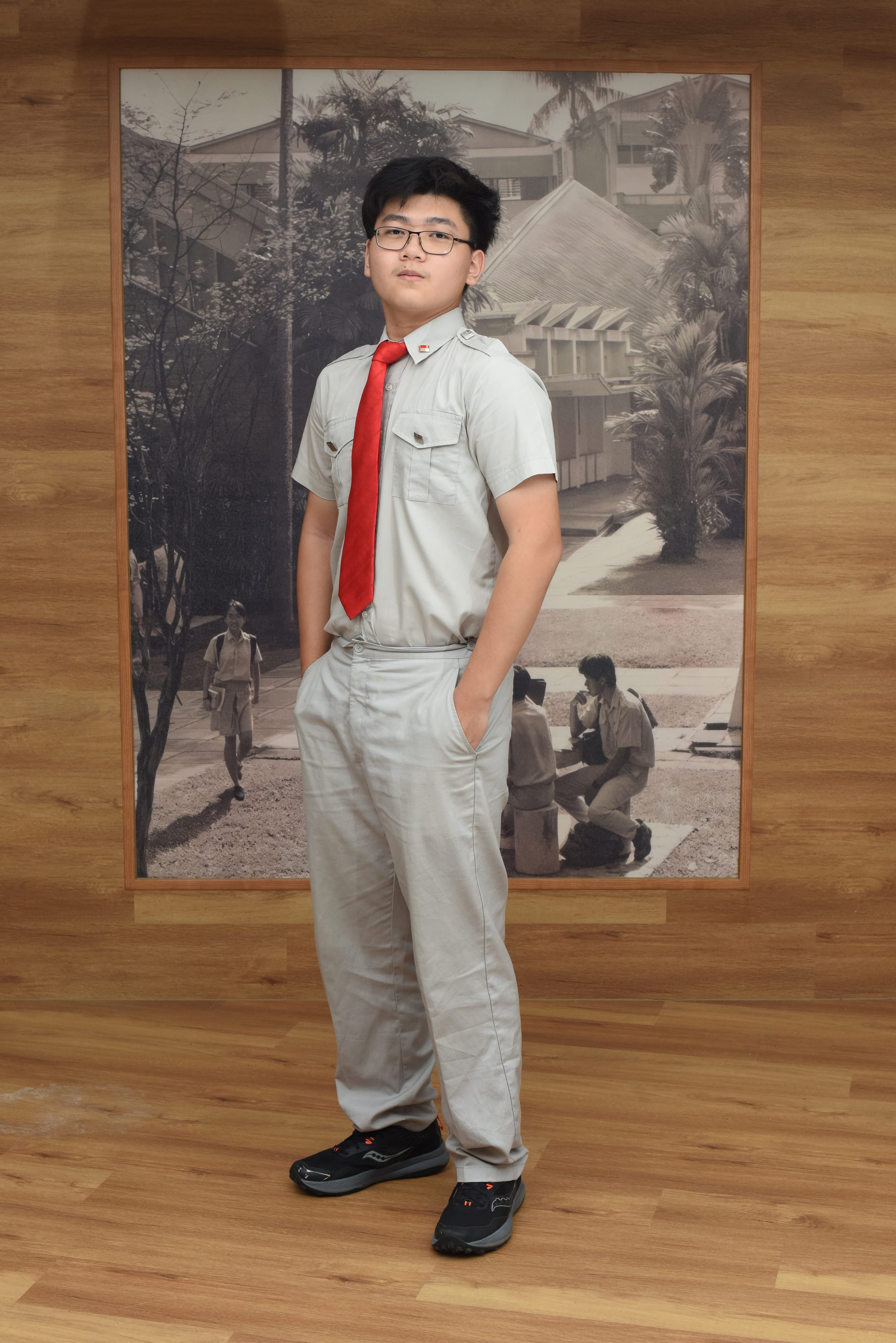 Young male student wearing glasses, gray uniform with red tie, standing on wood floor before a vintage black-and-white school photo.