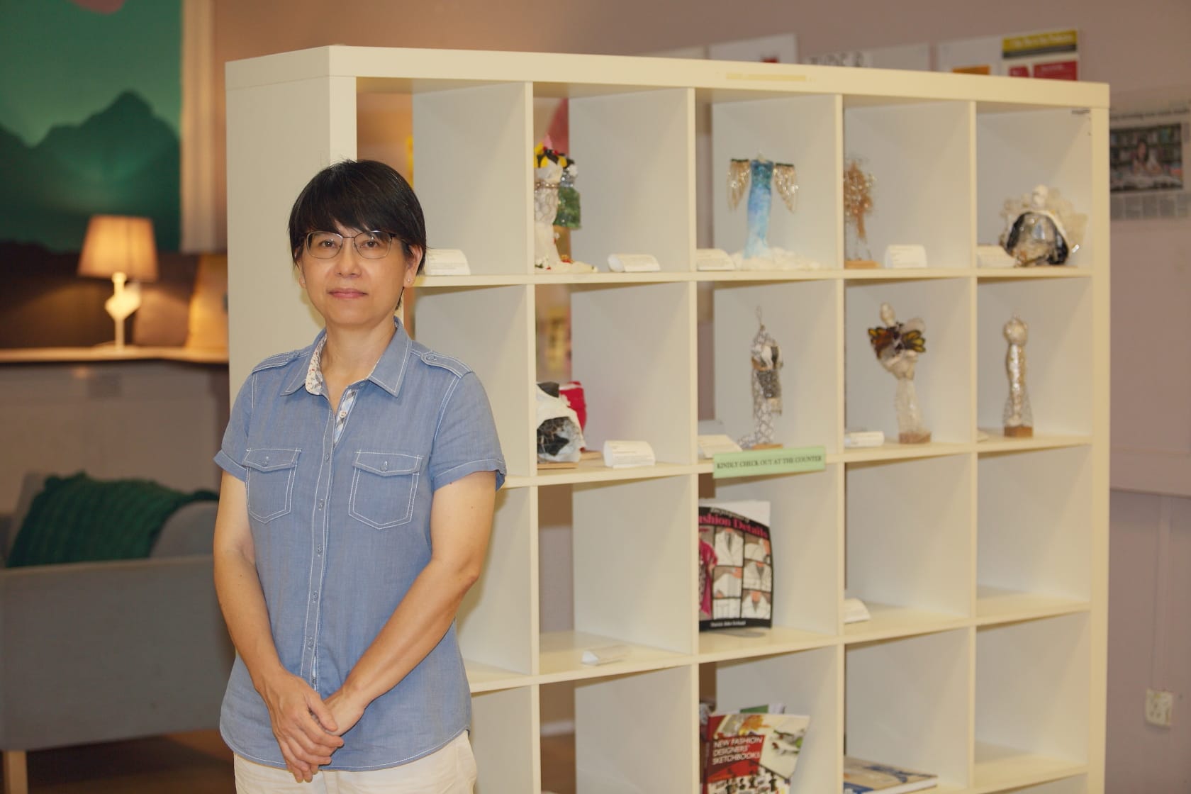 Woman with glasses in a blue shirt stands before a white cubby shelf displaying small figurines and books.