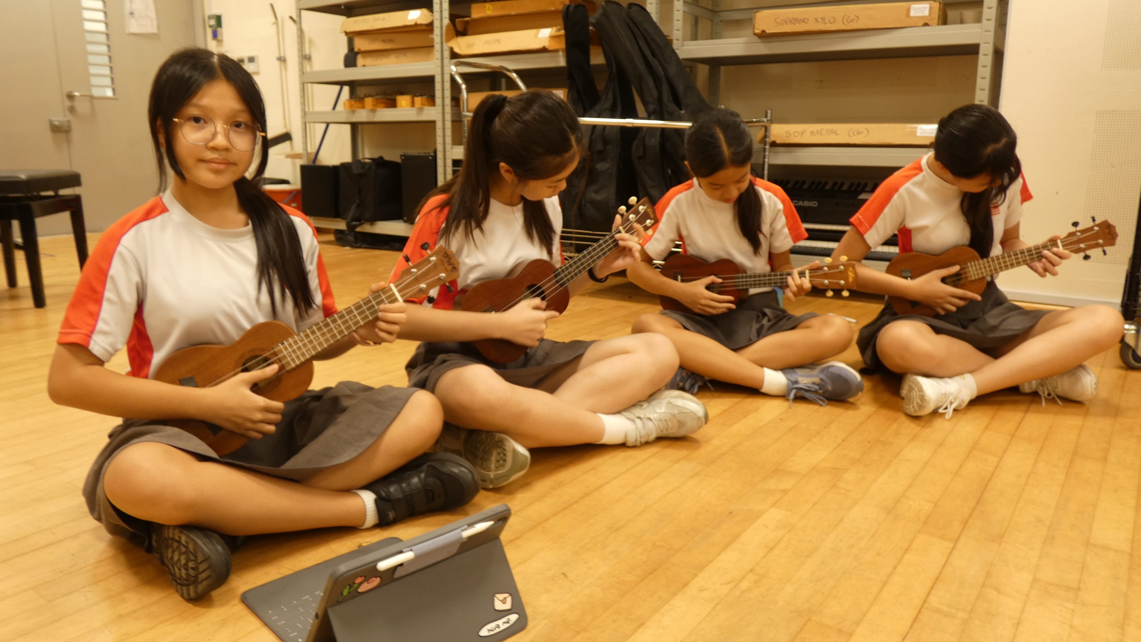 Four students sit on a wooden floor playing ukuleles in a music room with shelves and cases behind.