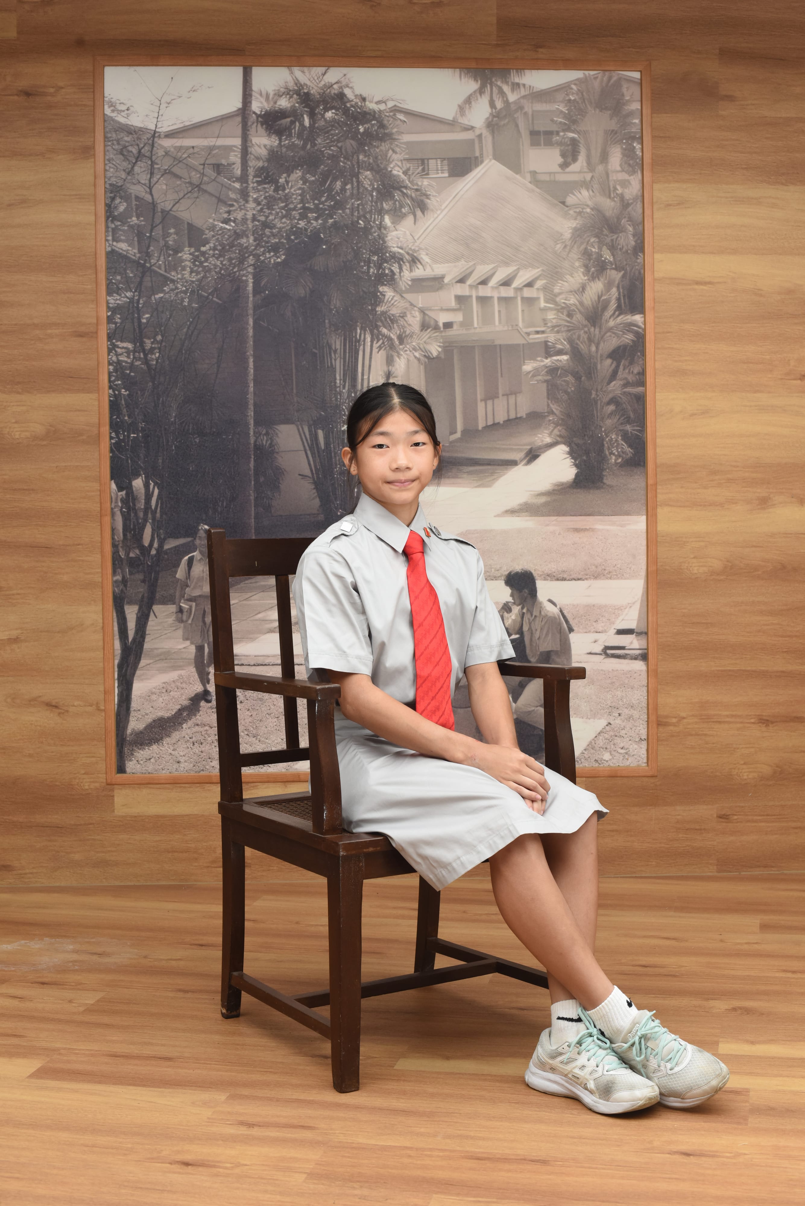 Young girl in a grey school uniform with a red tie sits on a wooden chair before a black and white photo mural.