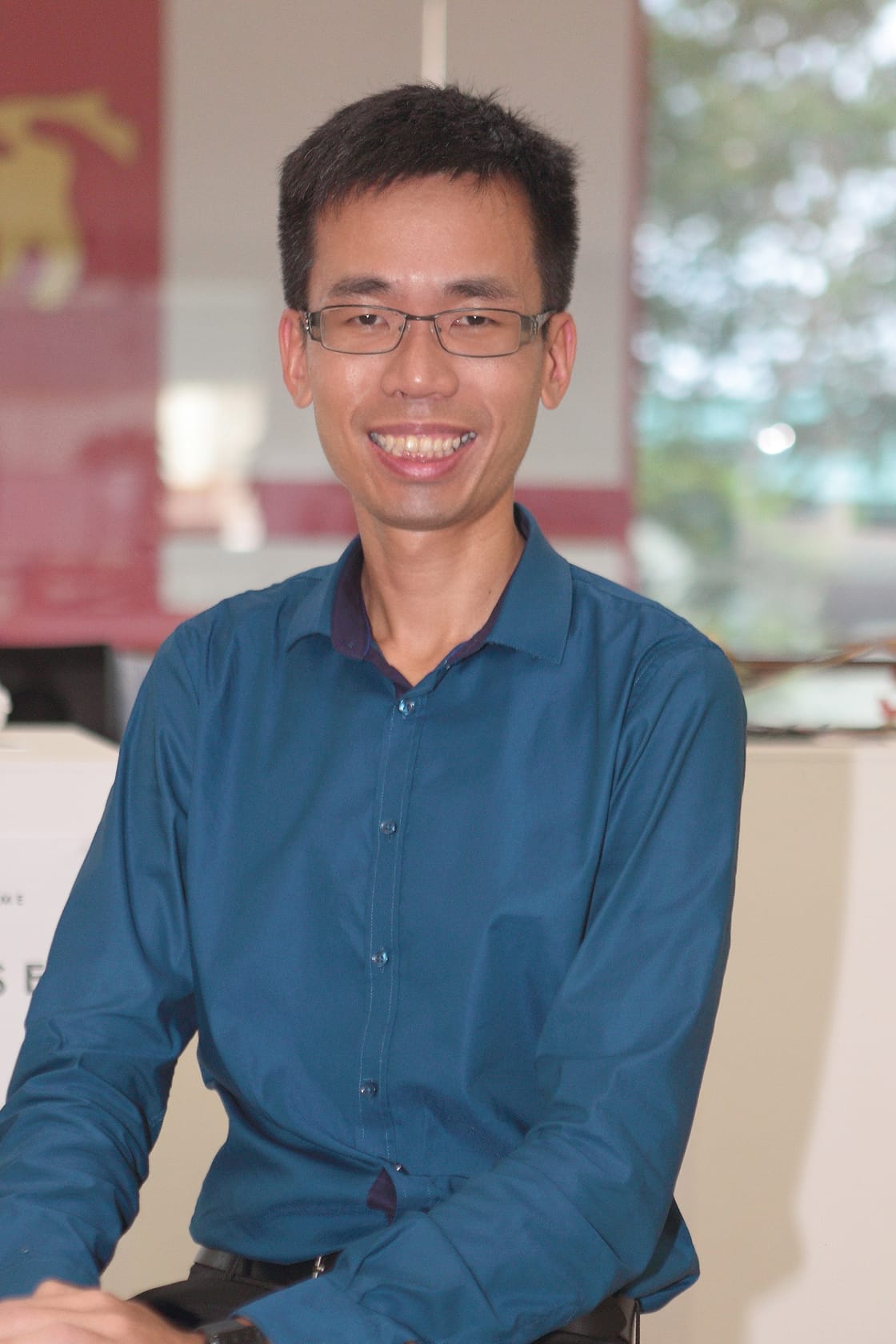 A smiling young man wearing glasses and a teal button-up shirt sits indoors with a blurred background.