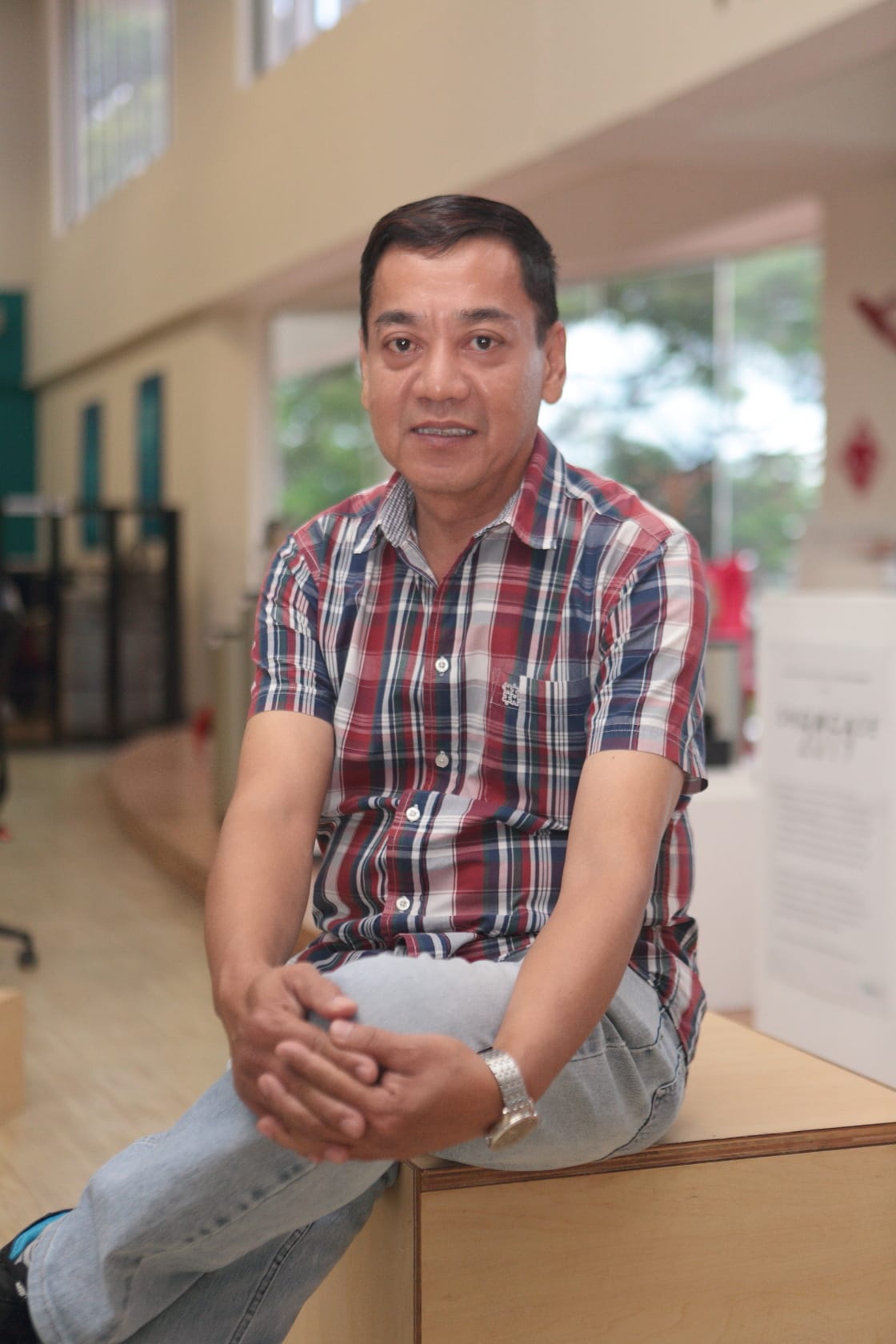 Middle-aged man in a red plaid shirt and jeans sits on a wooden box, smiling, inside a bright building.
