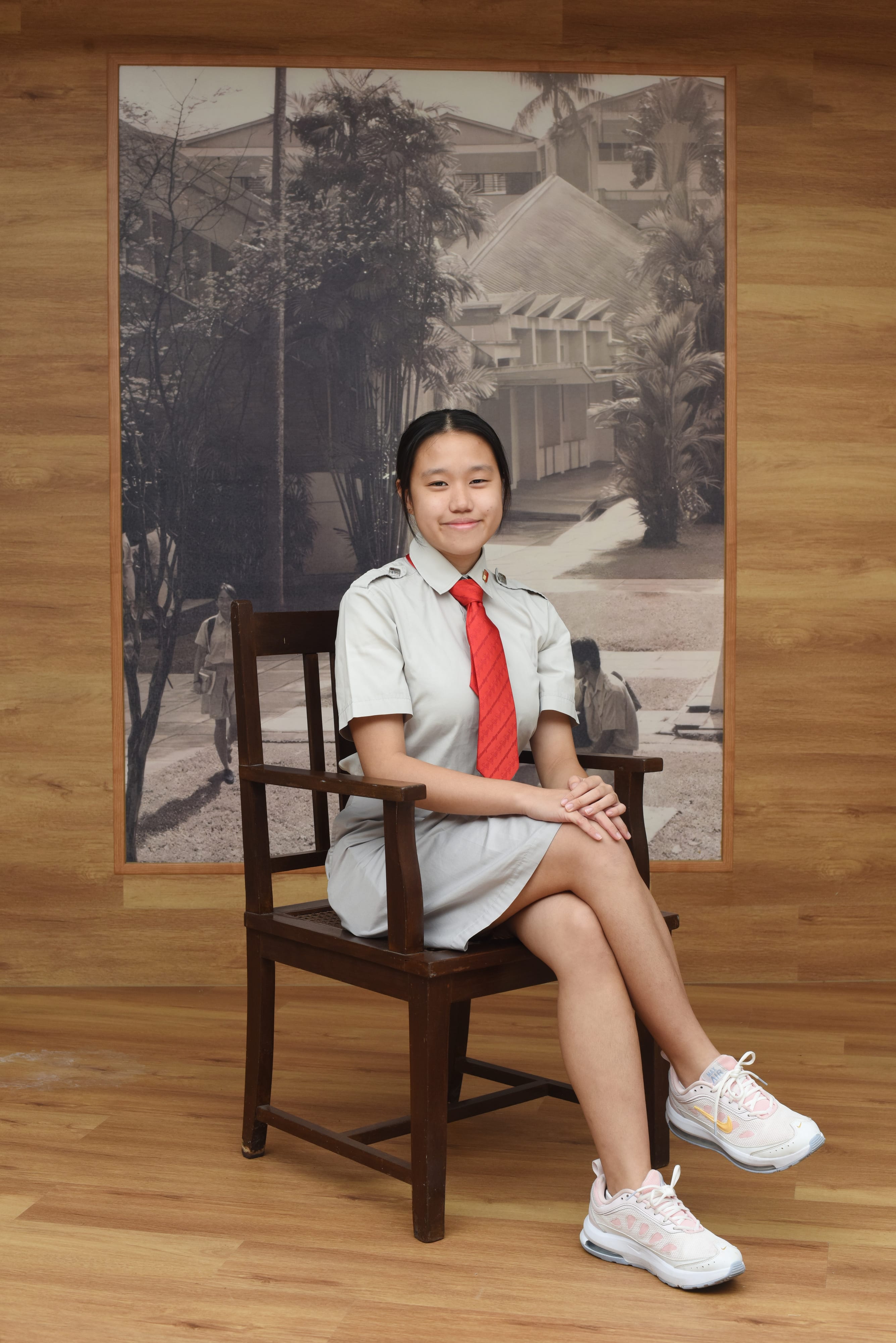 Young girl in a gray school uniform with a red tie sits on a dark wooden chair, smiling, in front of a black and white photo mural.