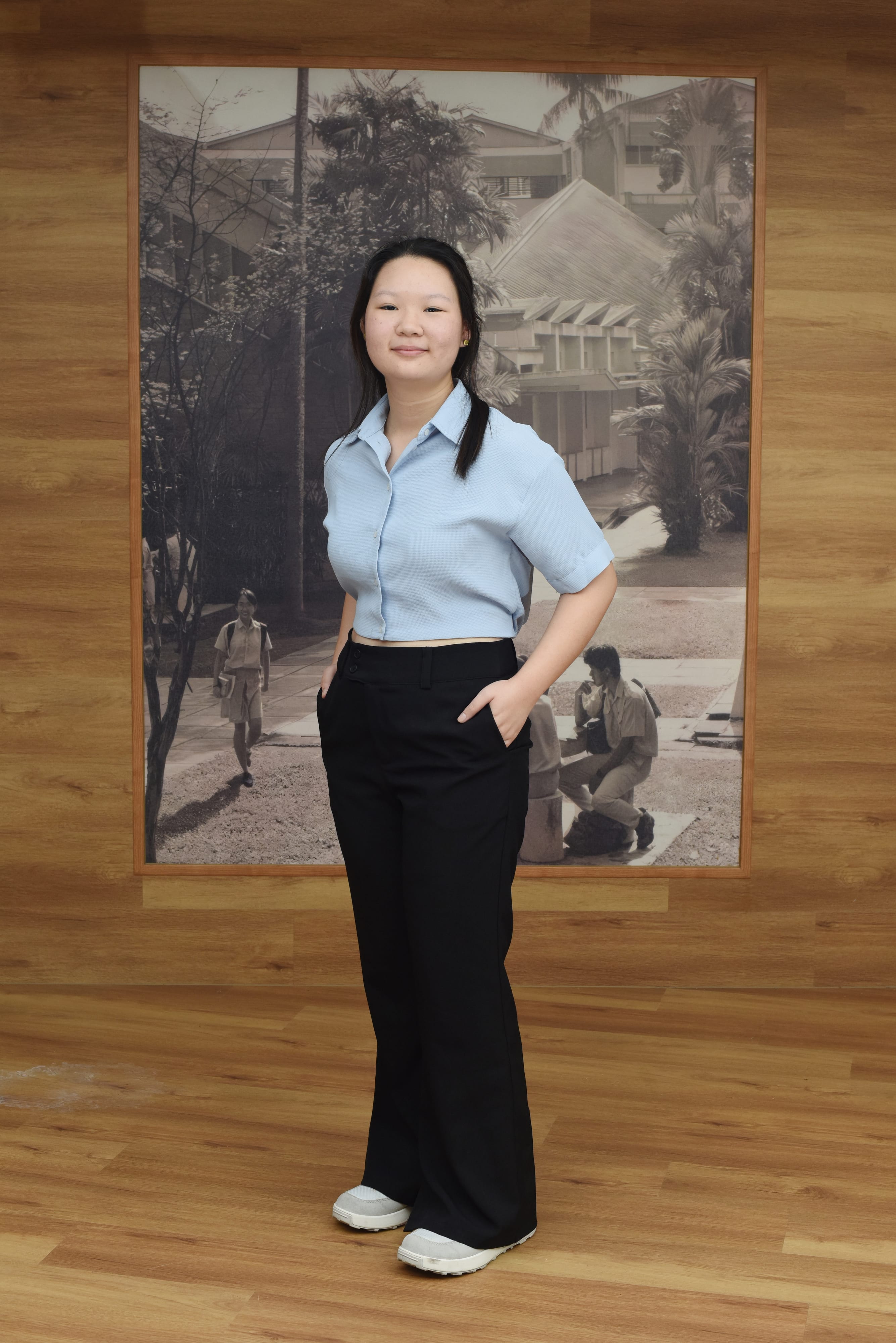 Young woman in a light blue shirt and black wide-leg pants standing before a large black and white photo on a wood wall.