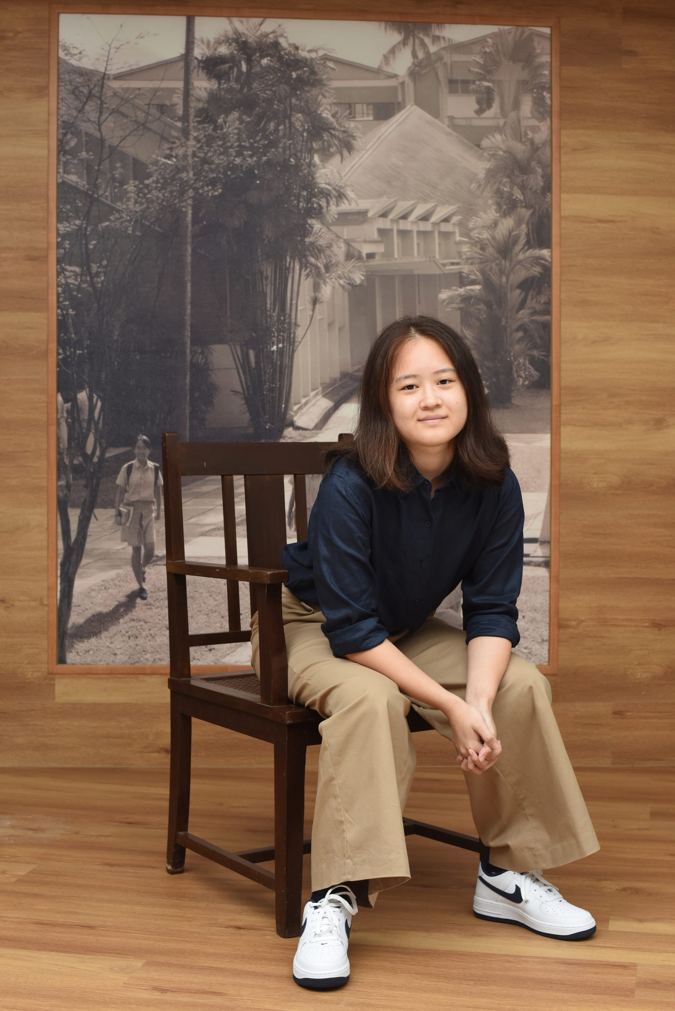 Young woman in navy shirt and khaki pants sits on a wooden chair before a black-and-white vintage building photo.