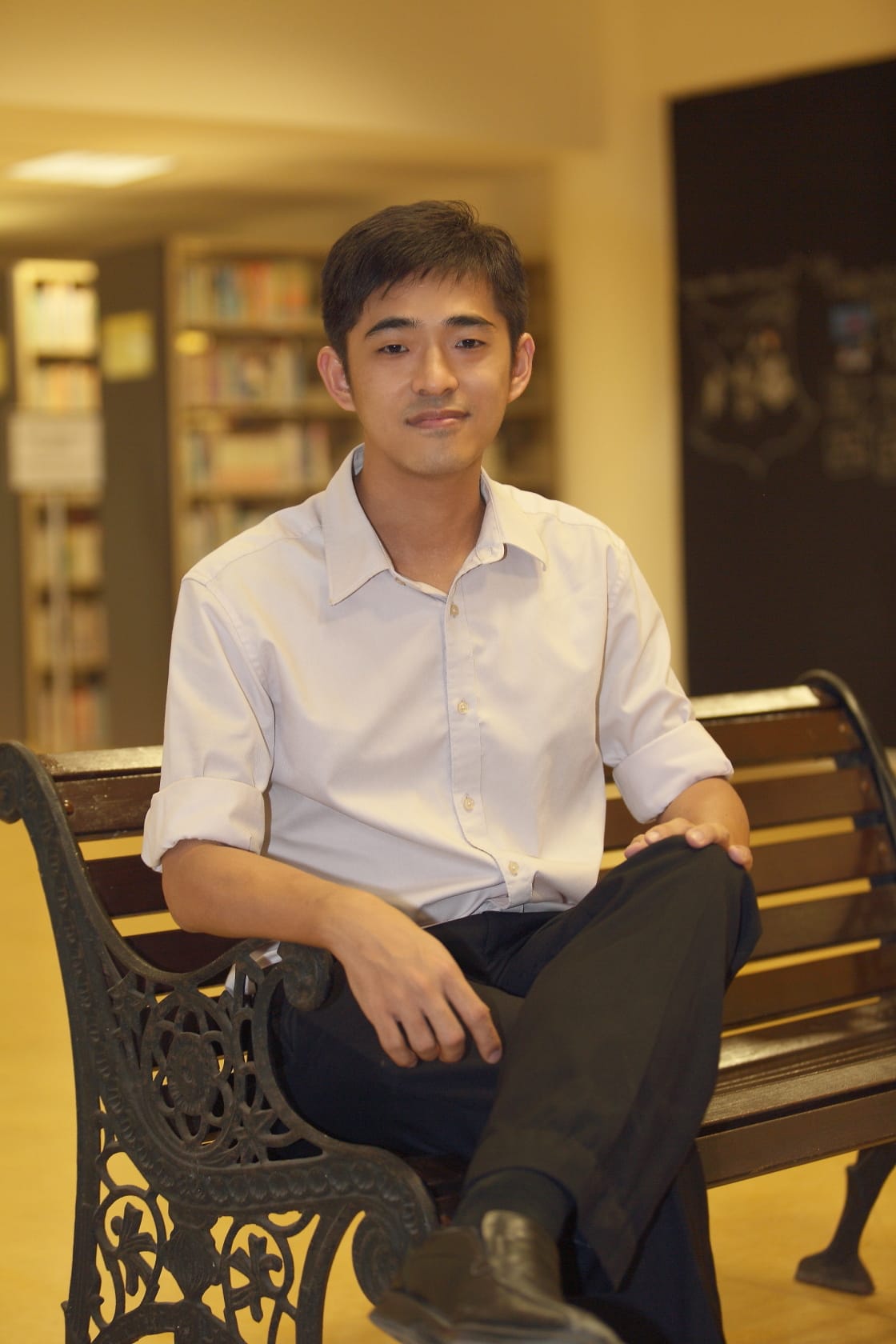 Young man in a white shirt and black pants sits on a wooden bench inside a library with bookshelves behind him.