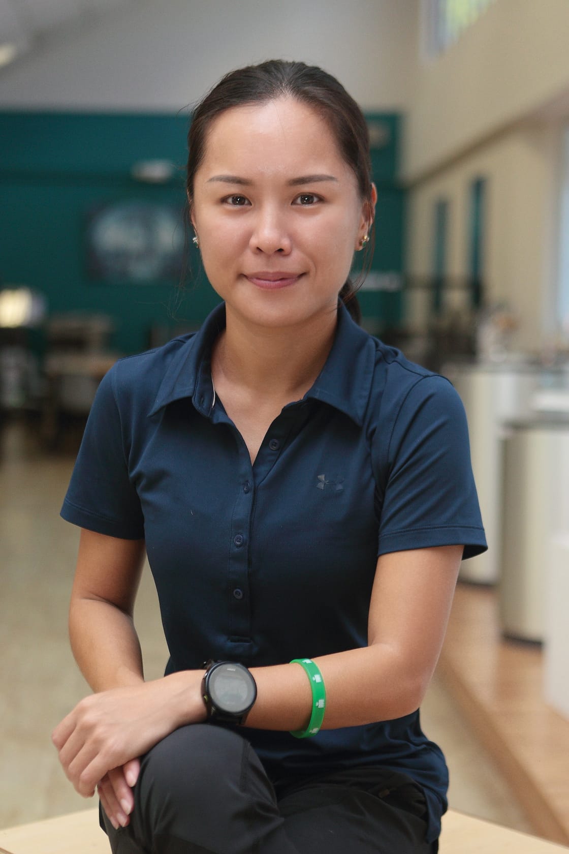 Woman with dark hair in a ponytail, wearing a navy polo shirt, black watch, and green wristband, sitting with arms crossed.