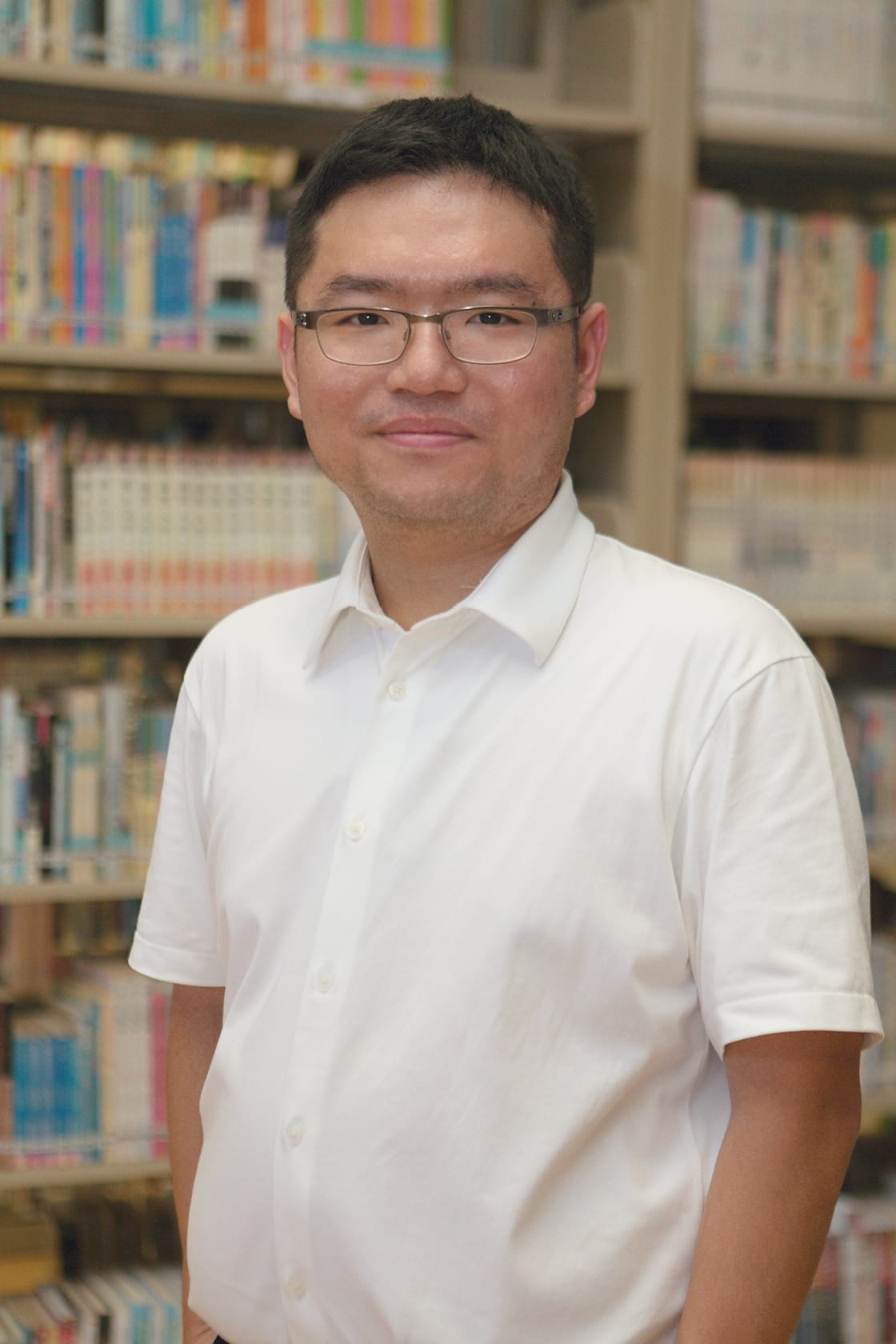 Man with glasses and white shirt smiling, standing in front of shelves filled with colorful books.