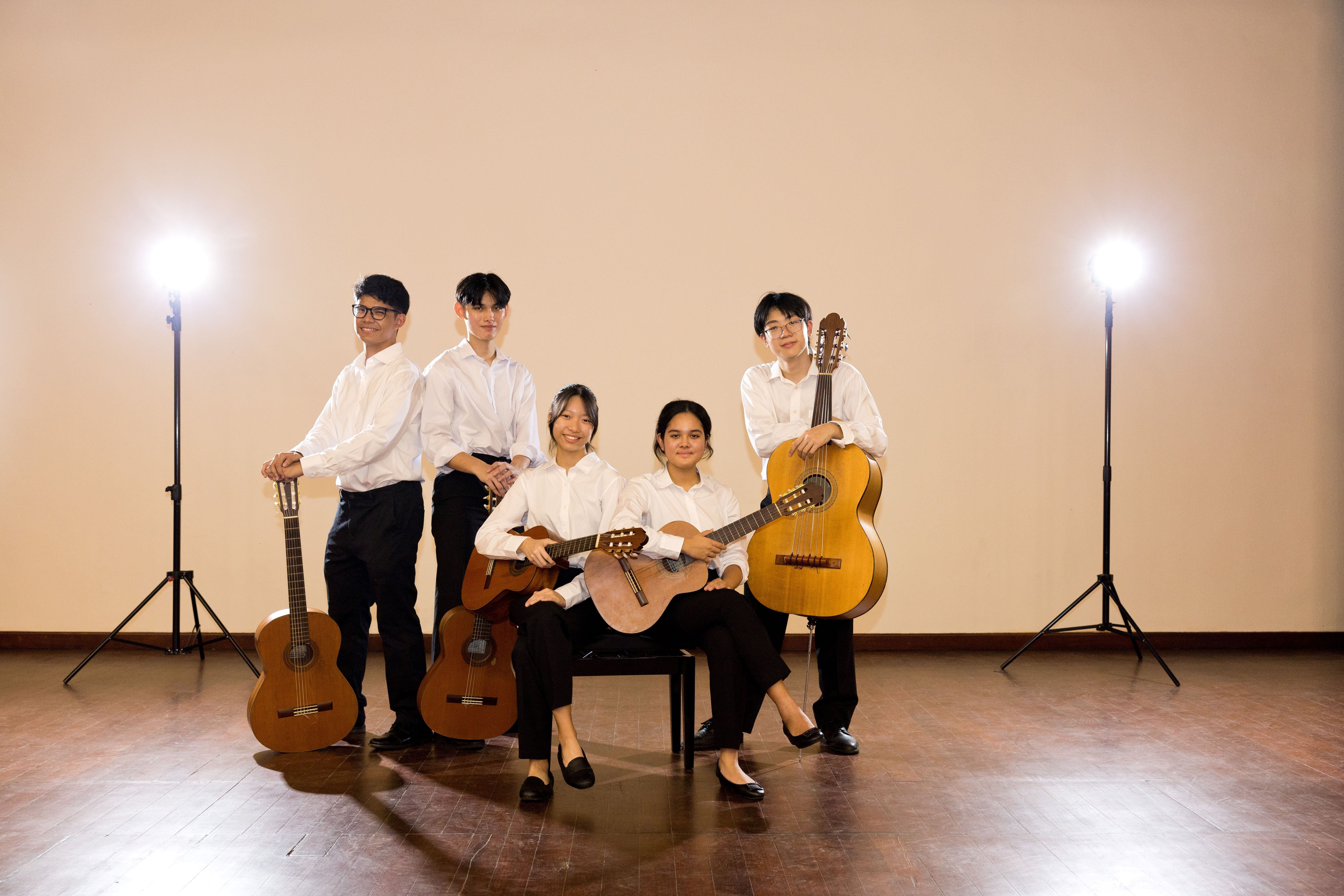 Five young musicians in white shirts and black pants pose with classical guitars under studio lights.