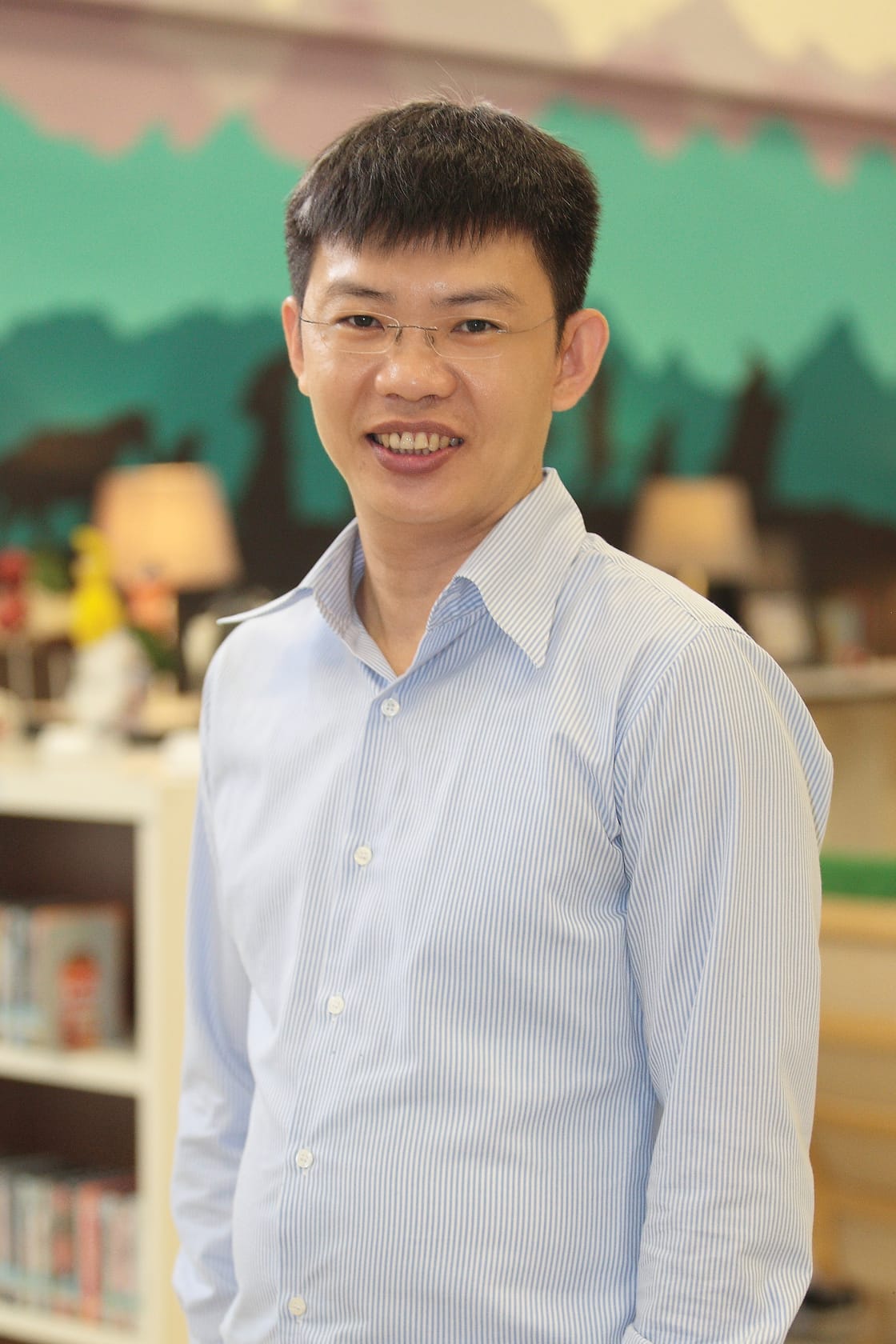 A smiling man with glasses wearing a light blue striped shirt stands in front of a colorful mural and bookshelves.