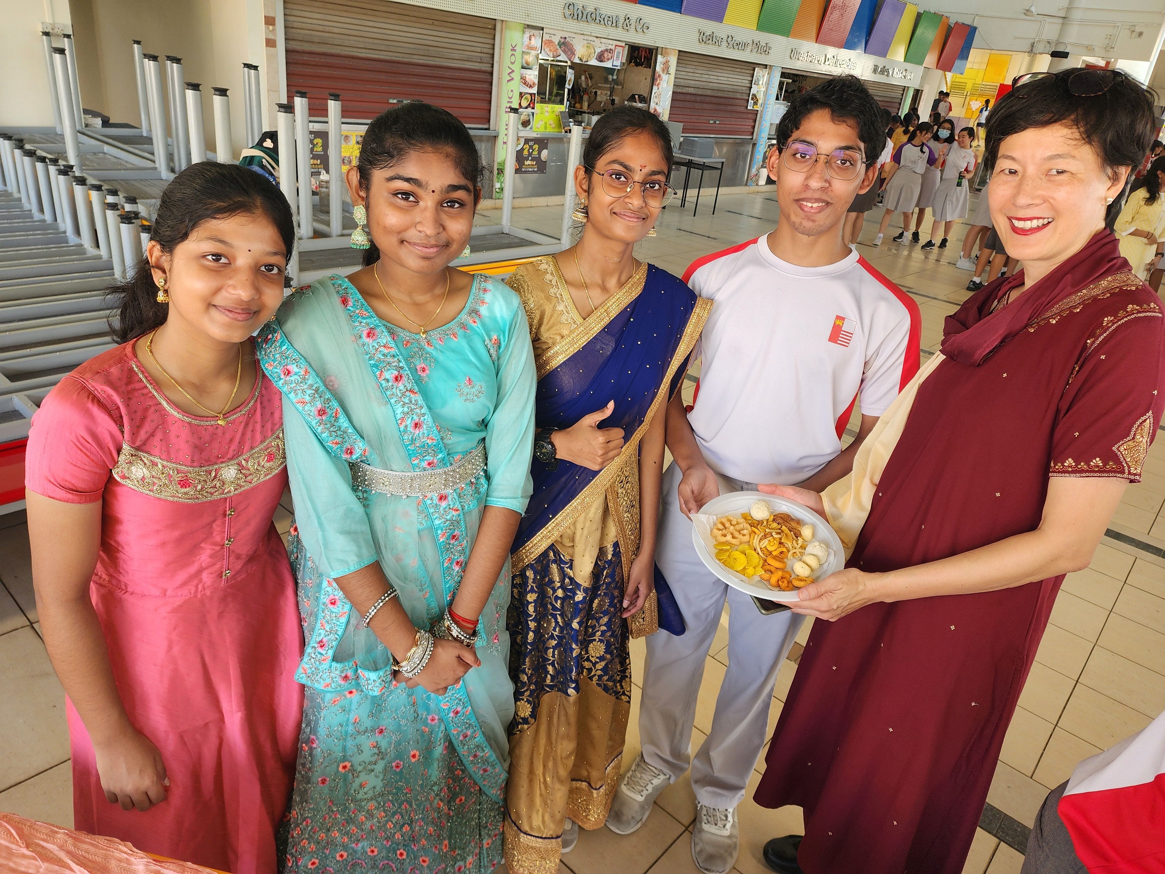 Five people smiling indoors, three in colorful Indian attire, one holding a plate of snacks, in a school canteen.