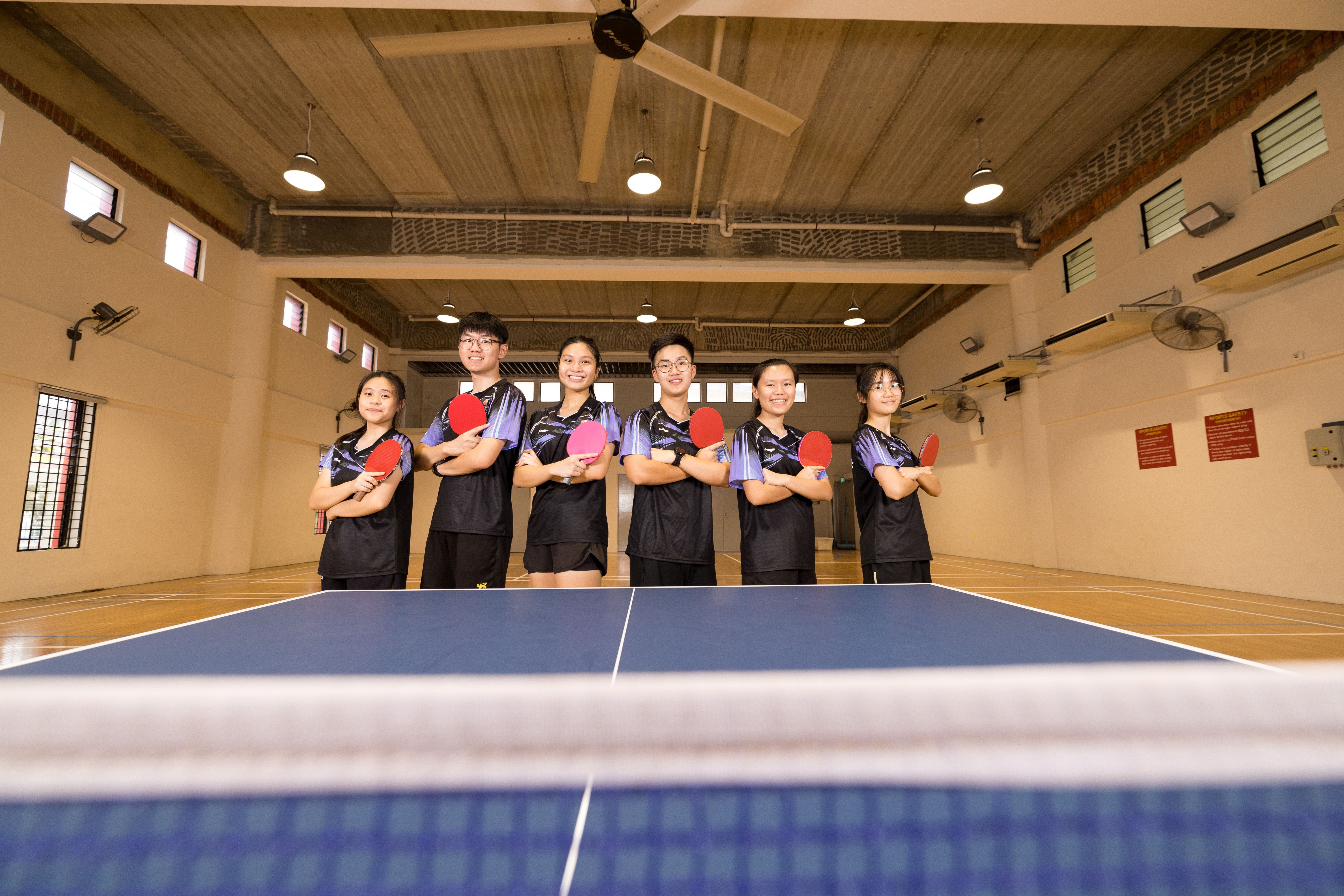 Six young table tennis players in matching uniforms hold paddles while posing behind a blue table indoors.