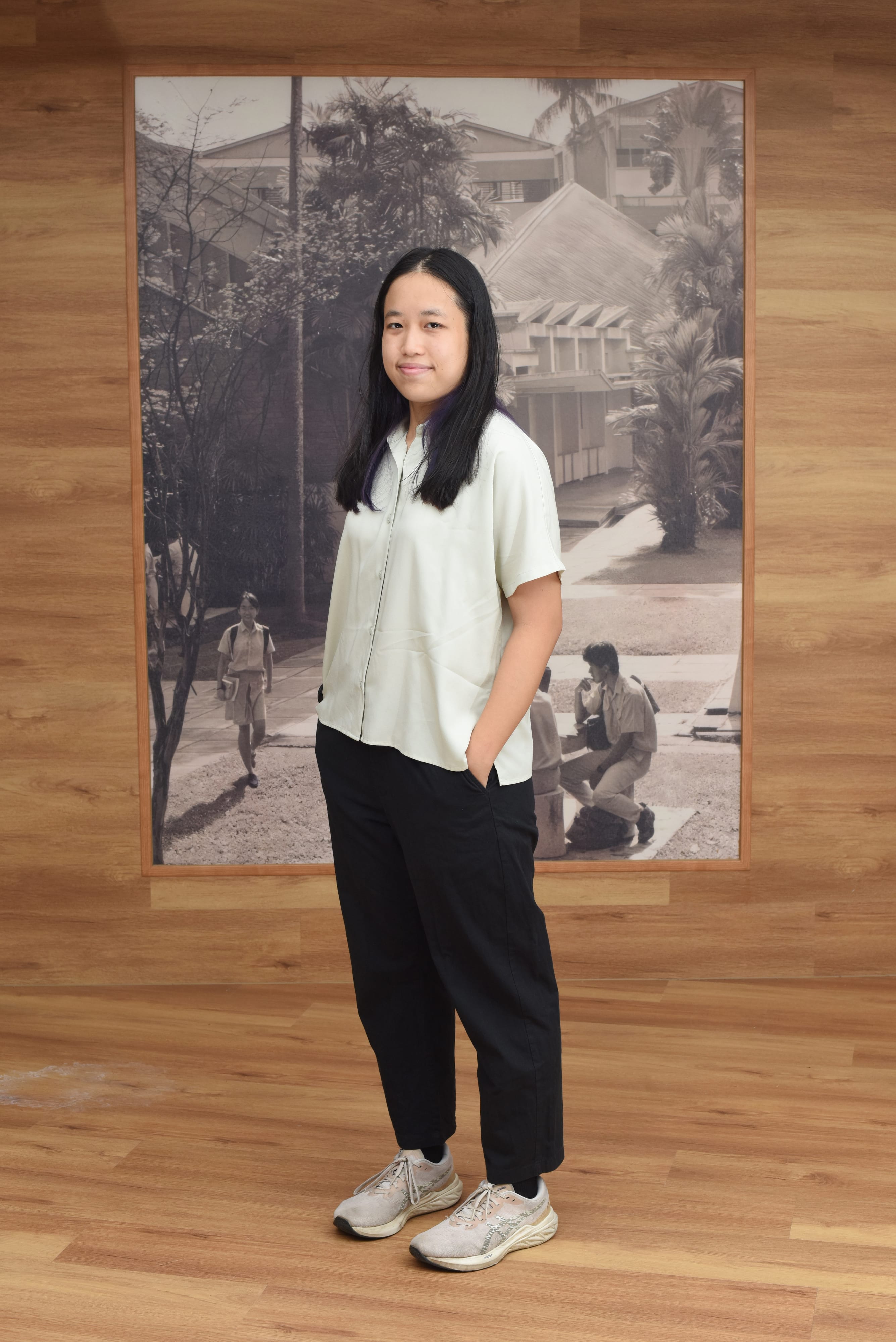 Young woman with long black hair in white shirt and black pants stands before a large black and white photo on a wood wall.