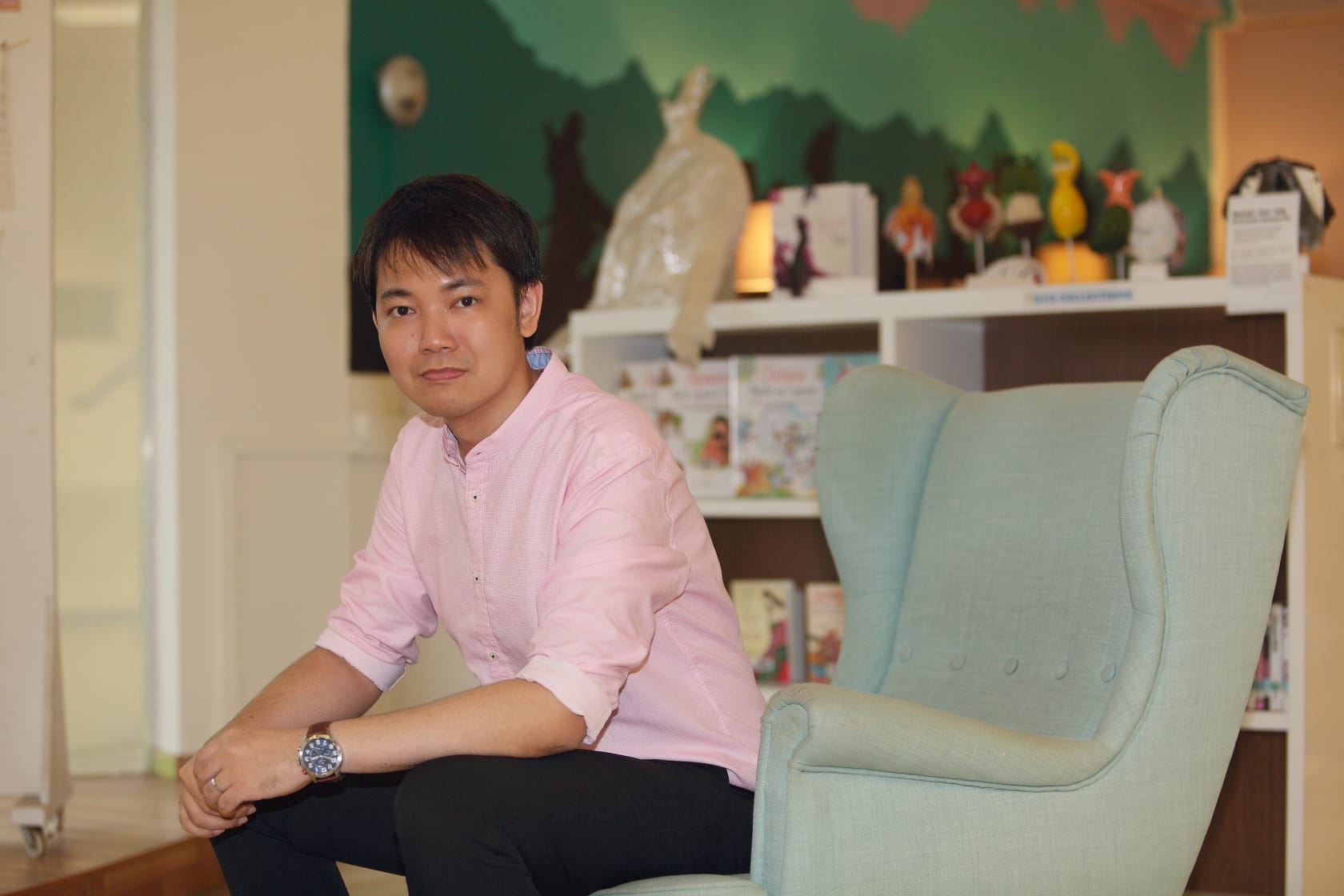 A young man in a pink shirt sits beside a light blue armchair in a colorful room with shelves and decorations.
