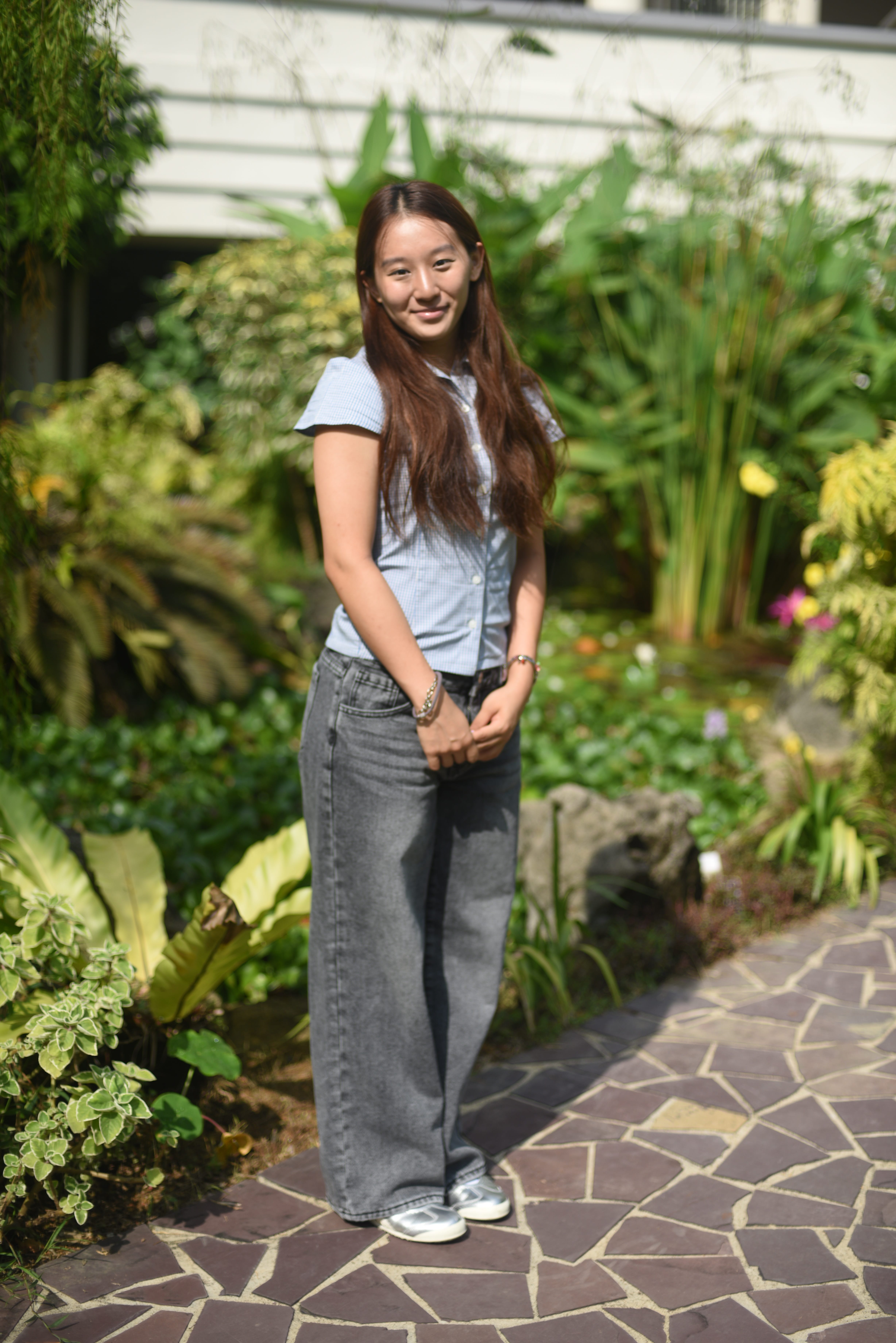 Woman with long hair in blue shirt and gray jeans standing on a stone path in a lush garden.
