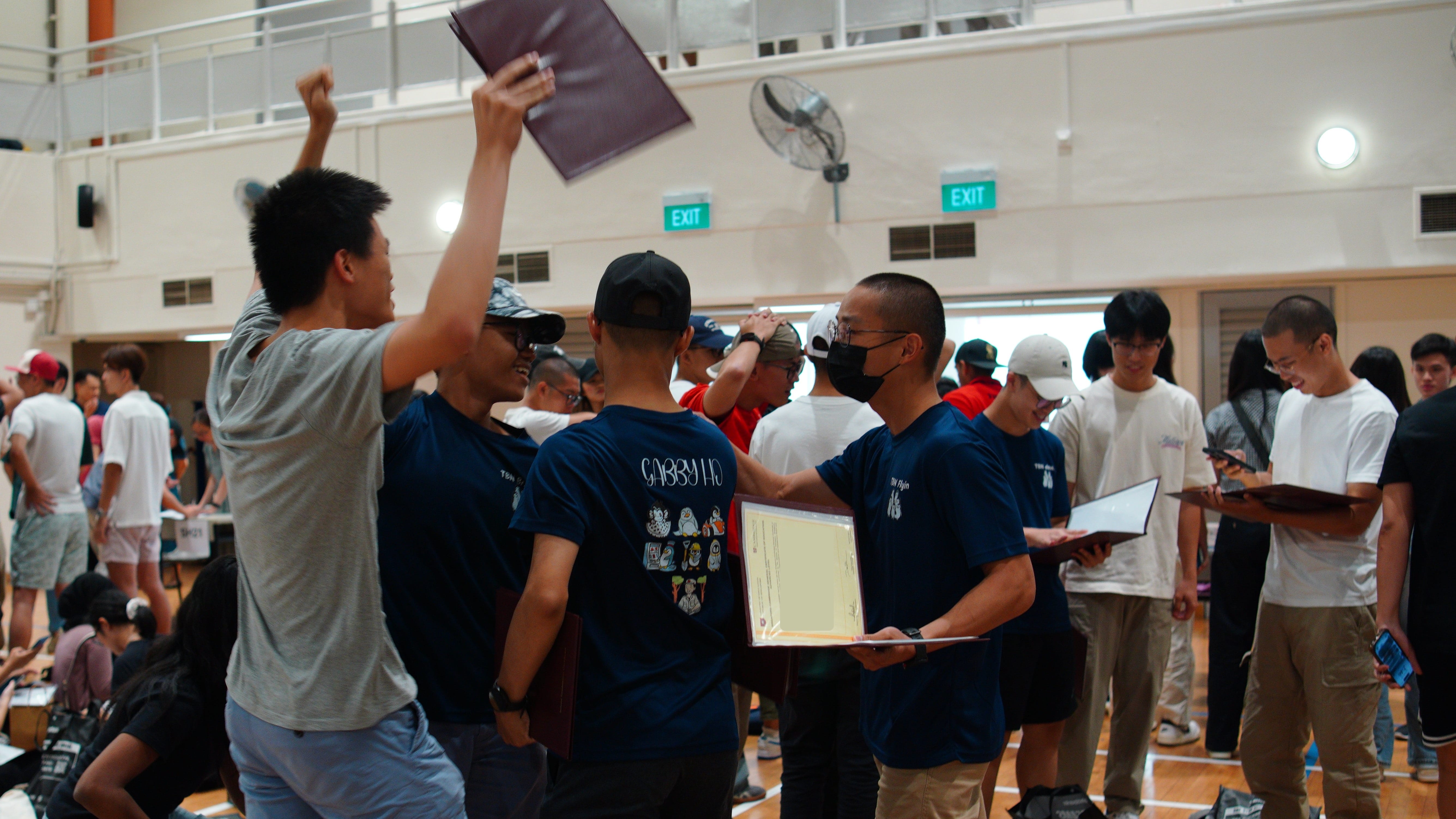 A young man raises a folder in celebration while another holds a certificate in a crowded indoor hall.