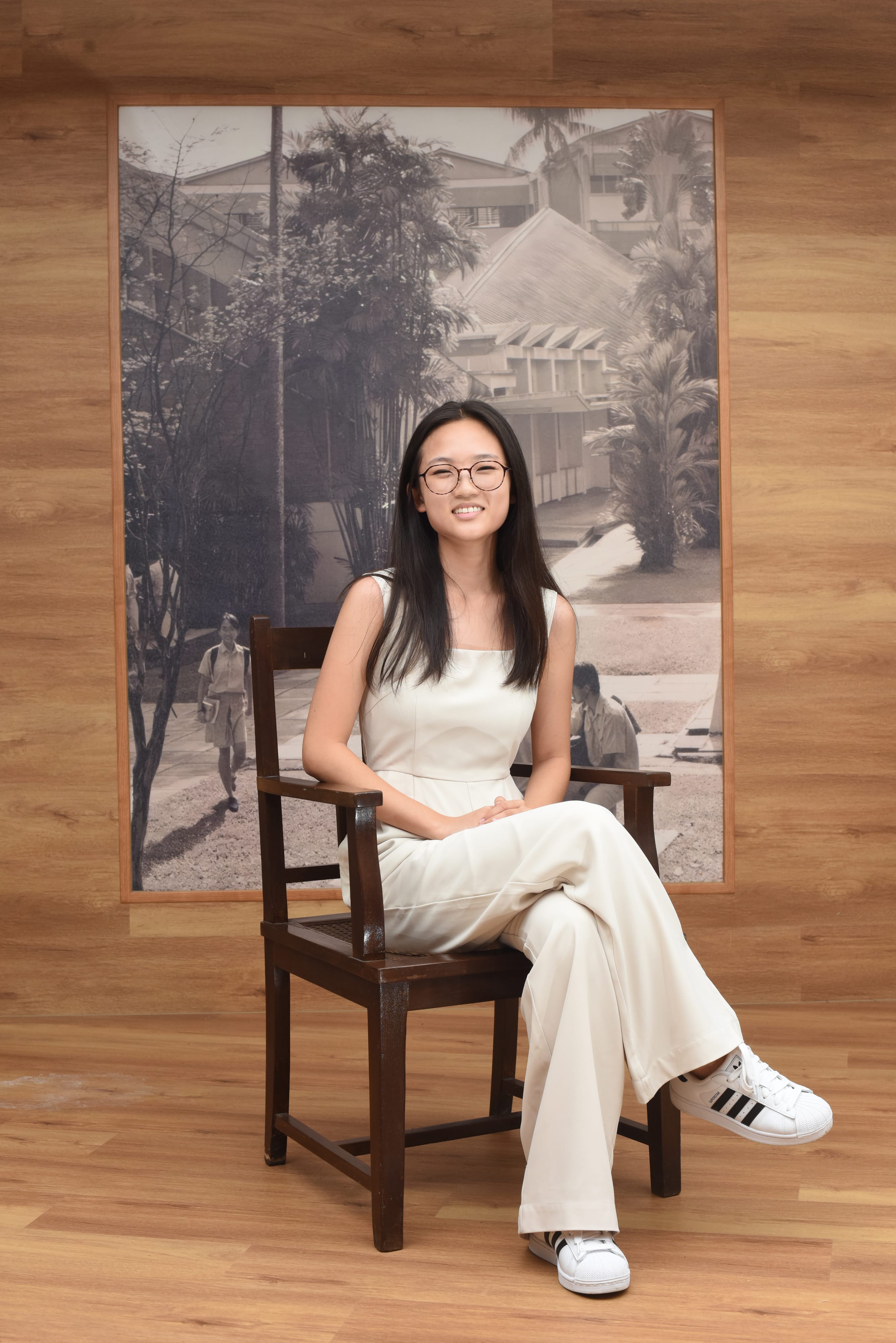 Young woman with glasses in white outfit sits on a wooden chair, smiling, before a large black-and-white photo of buildings.