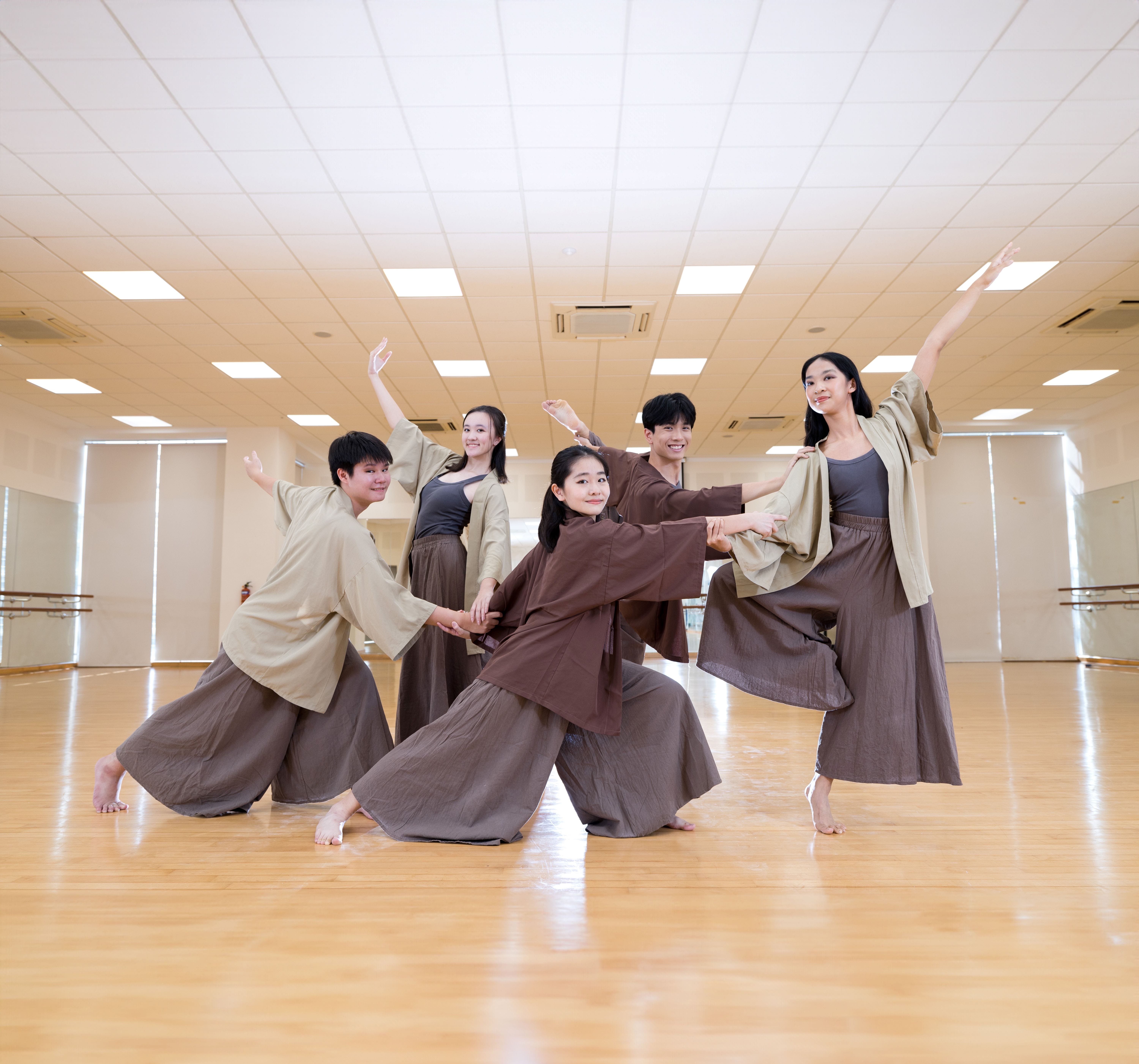 Five dancers in brown and tan flowing garments perform a coordinated pose in a mirrored dance studio.