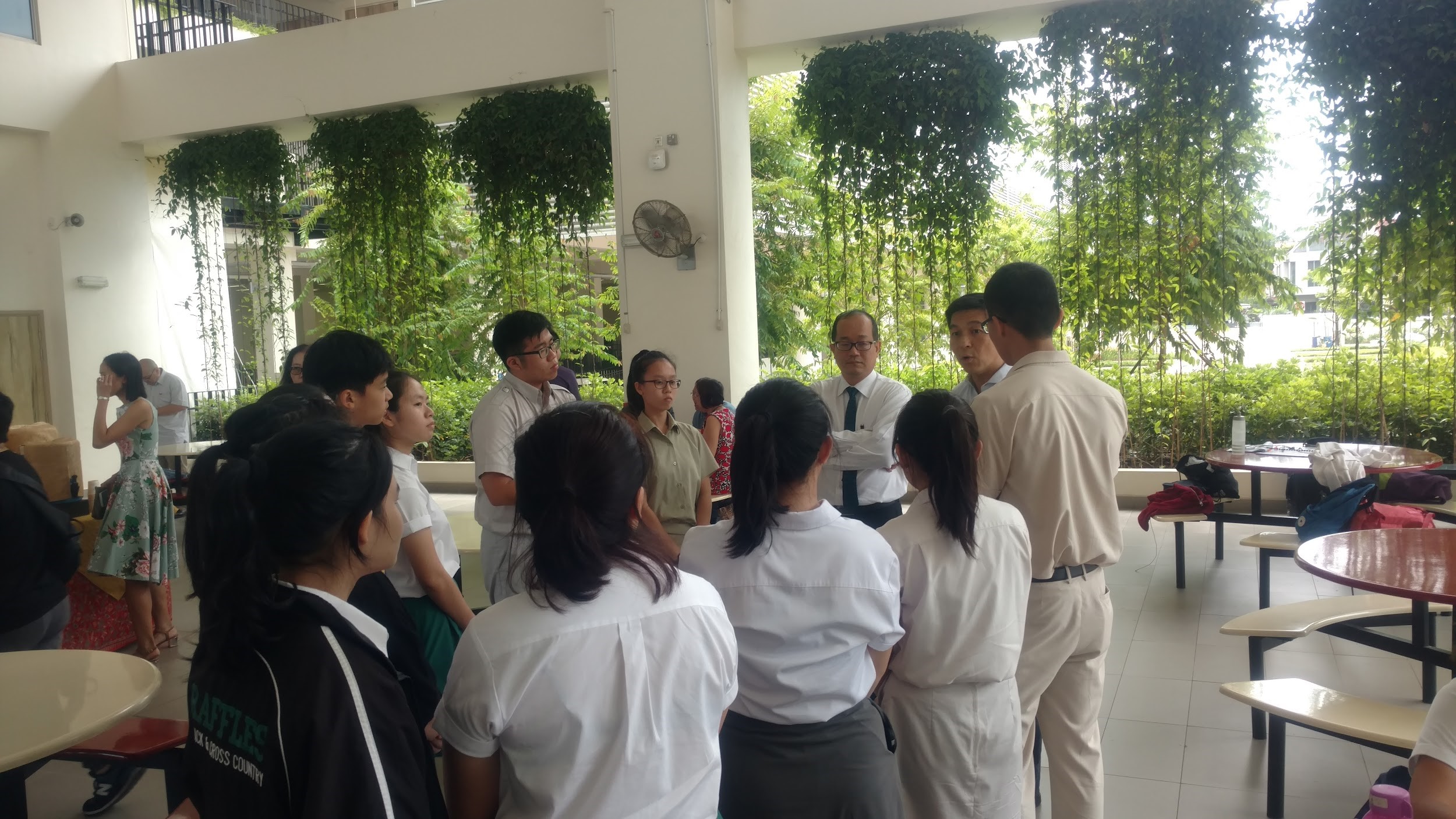 A group of students in white uniforms gather around two adults in a open hall with hanging green plants.