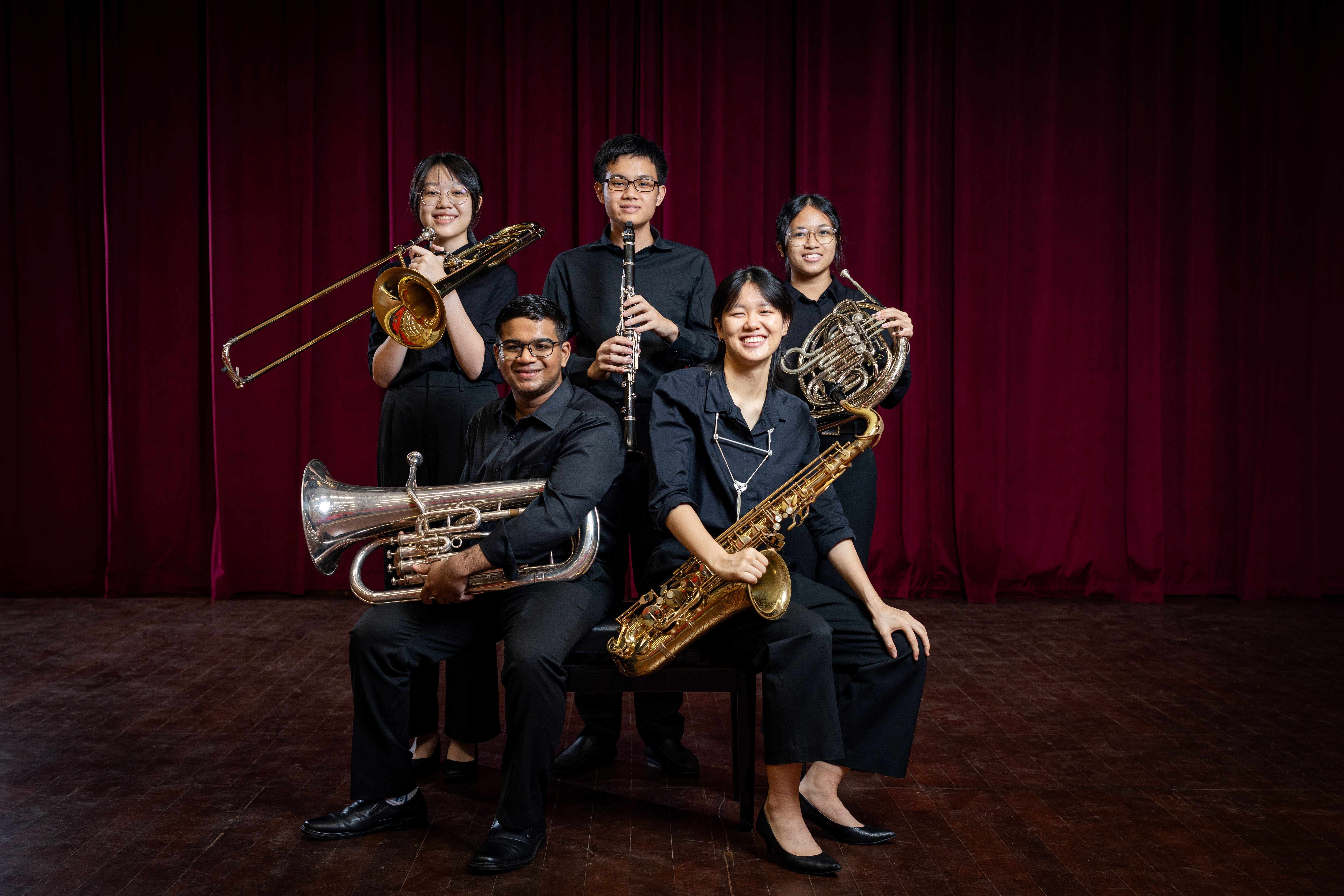 Five young musicians in black attire pose with brass and woodwind instruments before a red curtain.