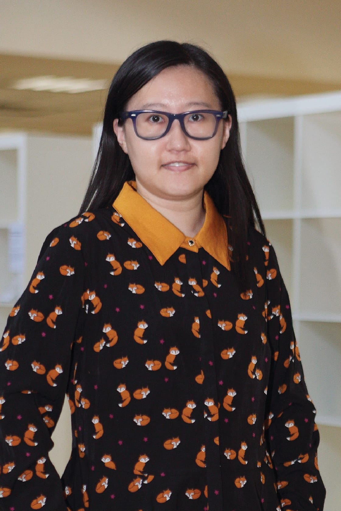 Woman with long black hair and blue glasses wearing a black shirt printed with orange foxes and a yellow collar.