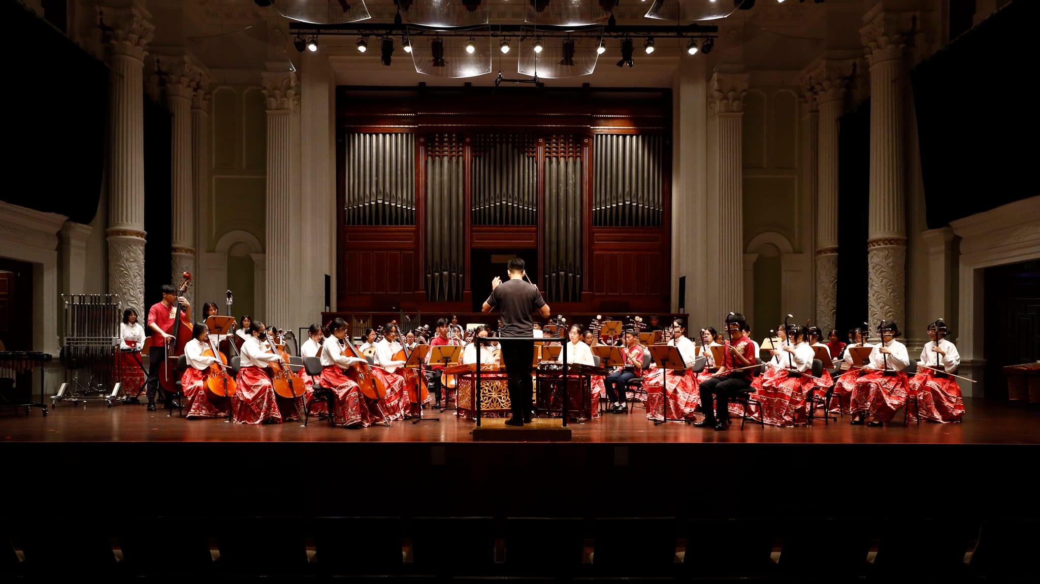 A conductor leads a large orchestra on a concert hall stage, with musicians wearing red and white traditional attire.