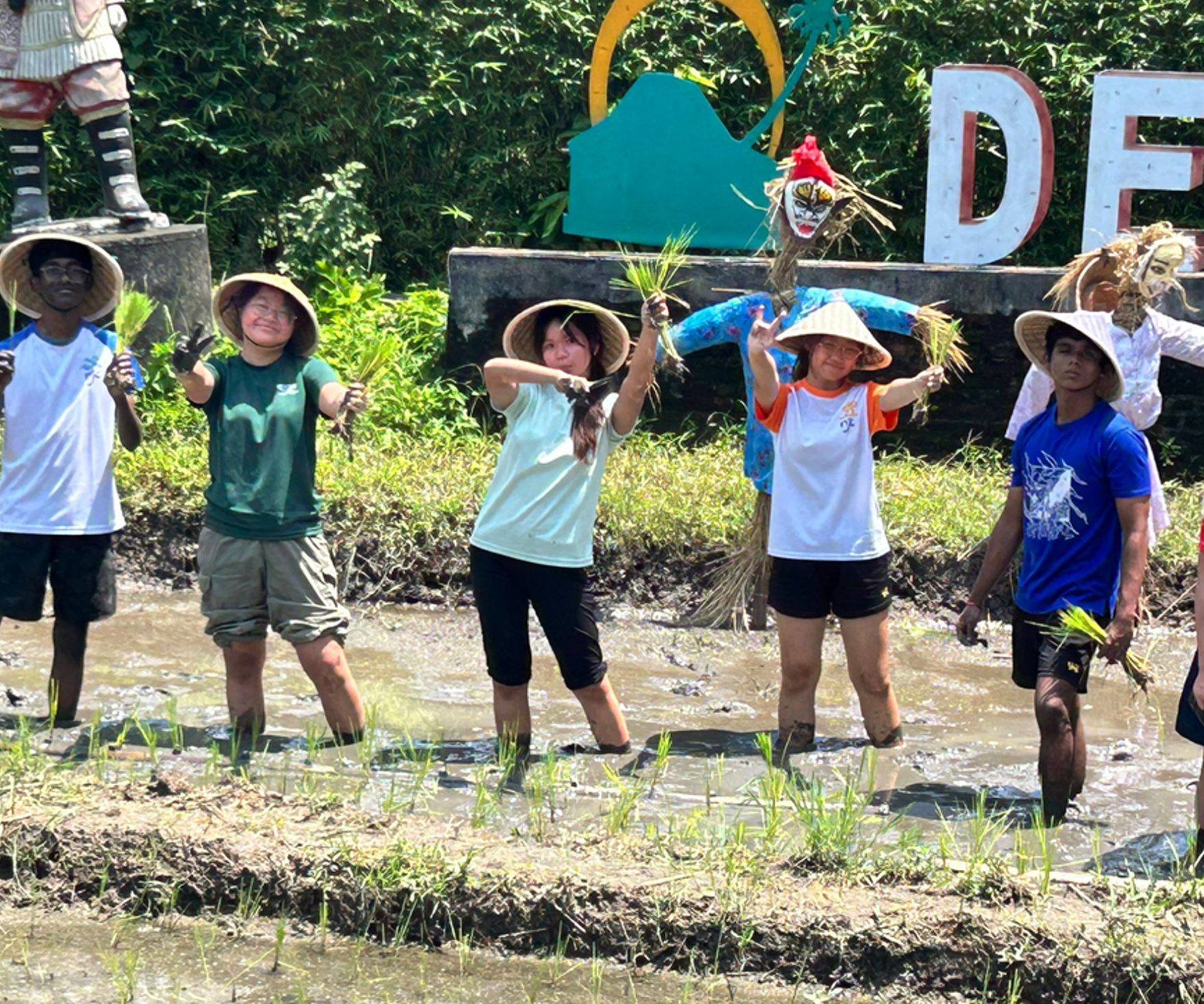 Five people in a muddy rice paddy holding rice seedlings, with scarecrows and large letters behind them.