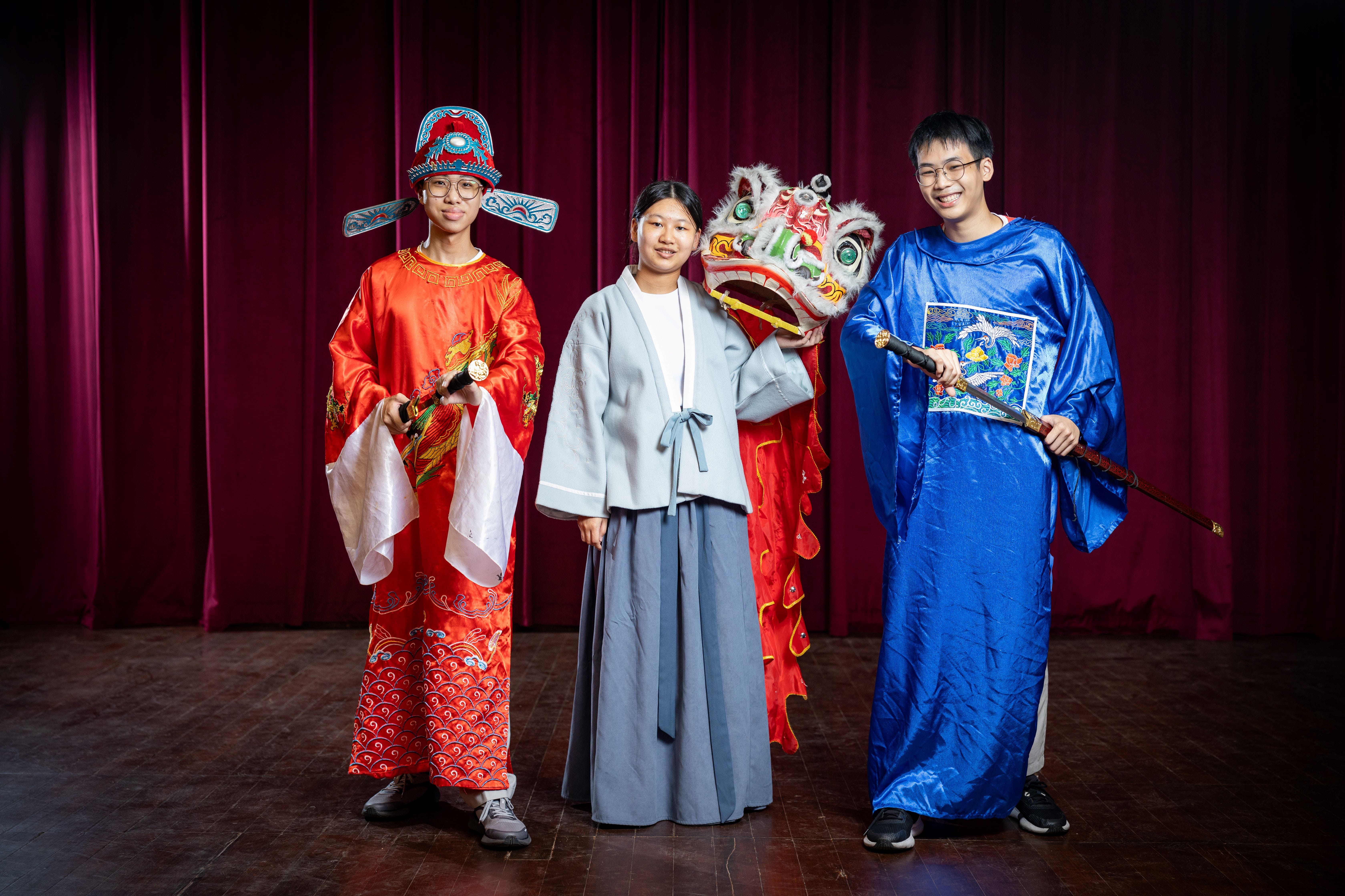 Three young people in traditional Chinese costumes pose on a stage, one holding a lion dance head, before a red curtain.