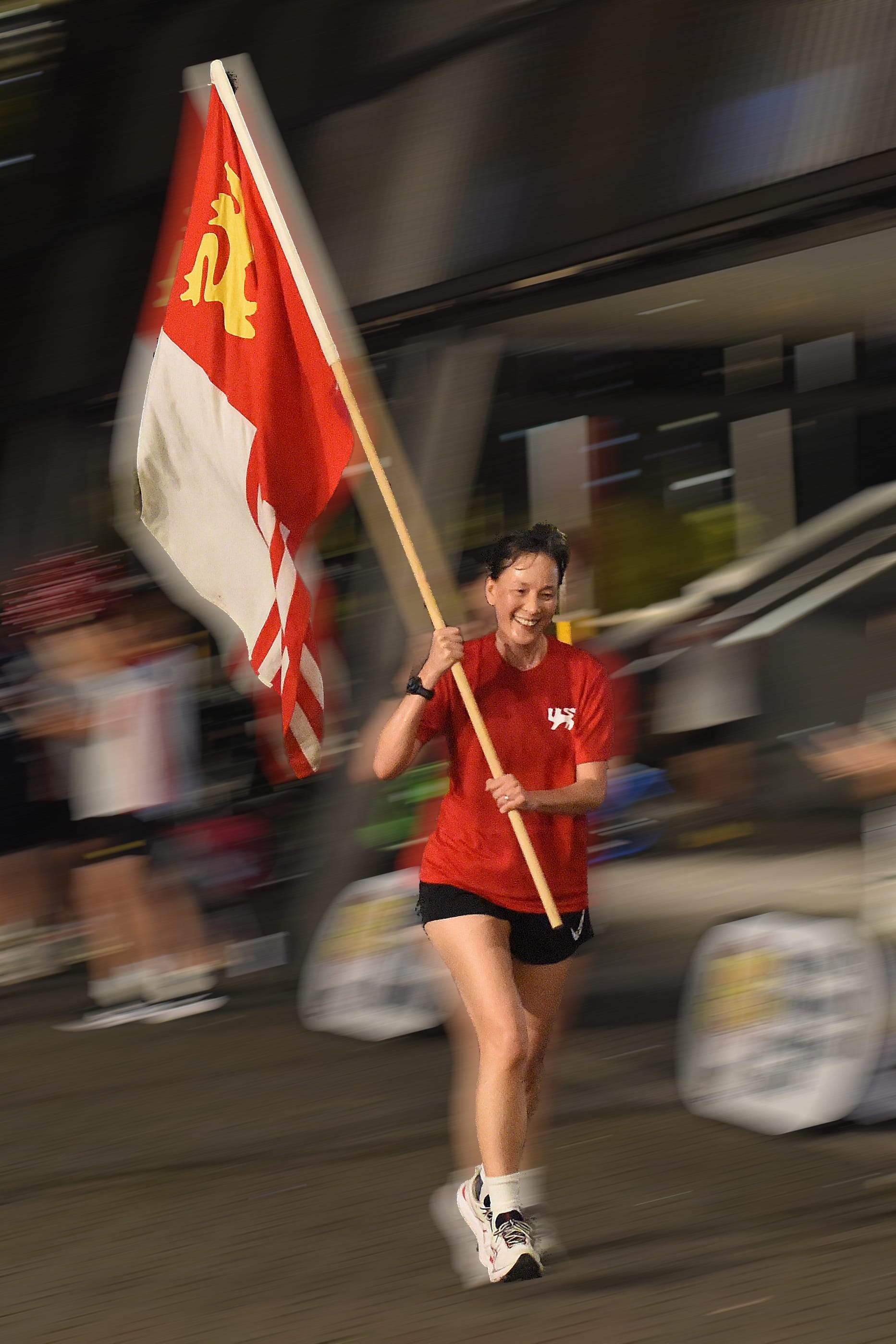 A smiling woman in a red shirt runs while carrying a red, white, and gold flag on a wooden pole.