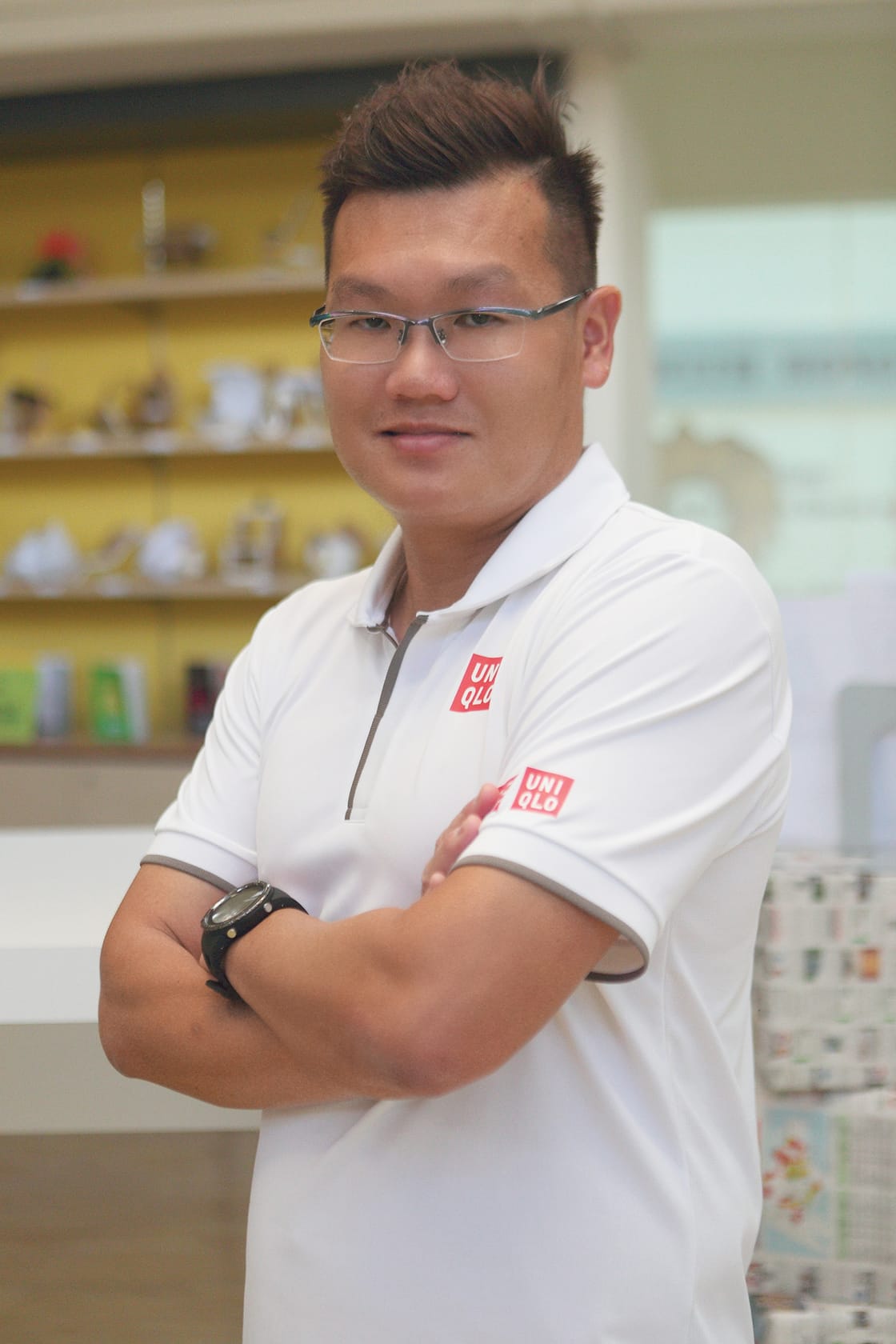 A young man with glasses and a black watch stands arms crossed, wearing a white Uniqlo polo shirt indoors.