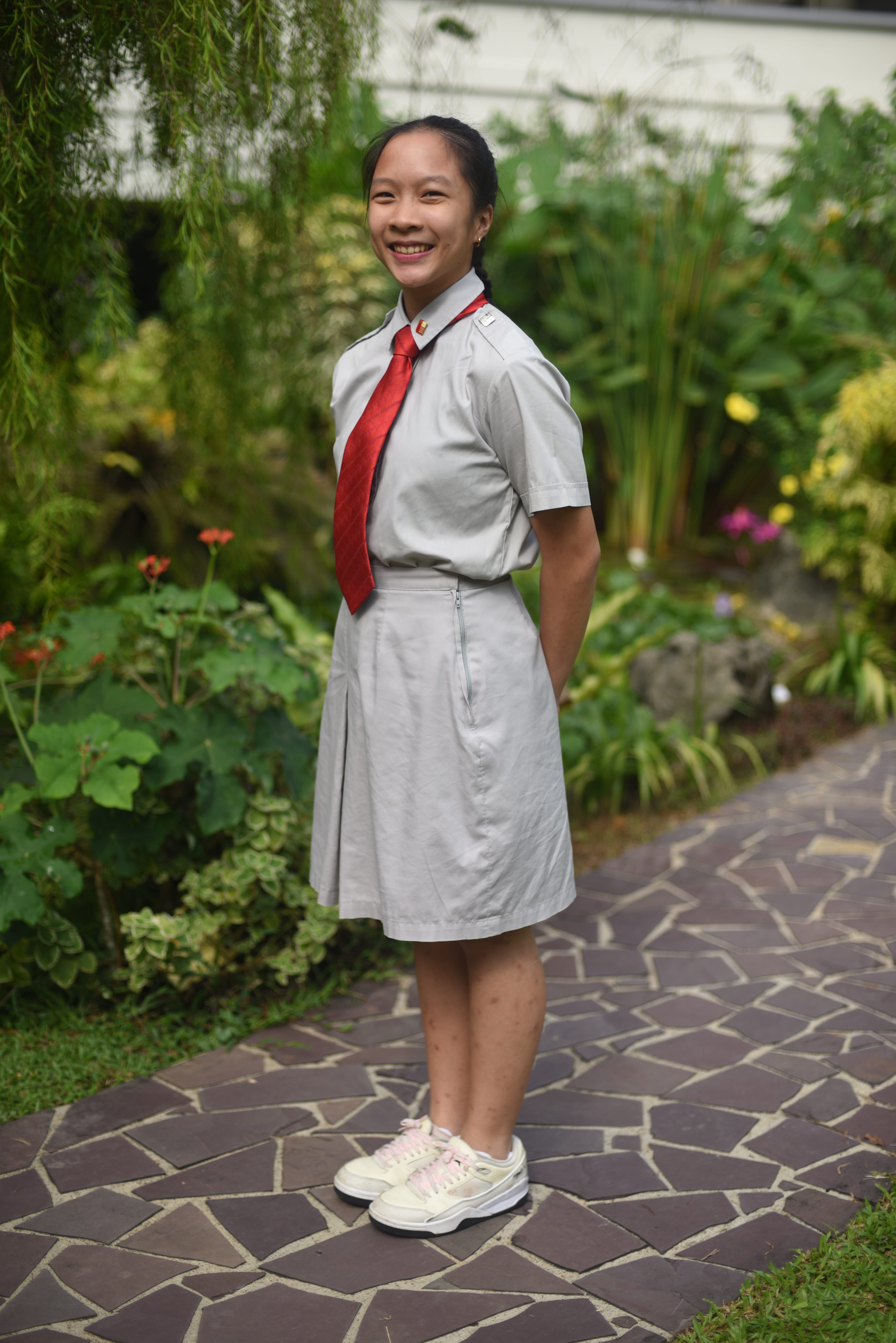 Smiling student in gray uniform with red tie stands on stone path in a lush garden.