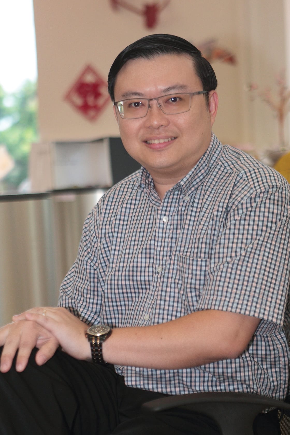 A smiling man with glasses and dark hair wearing a plaid shirt and watch, seated with hands clasped.