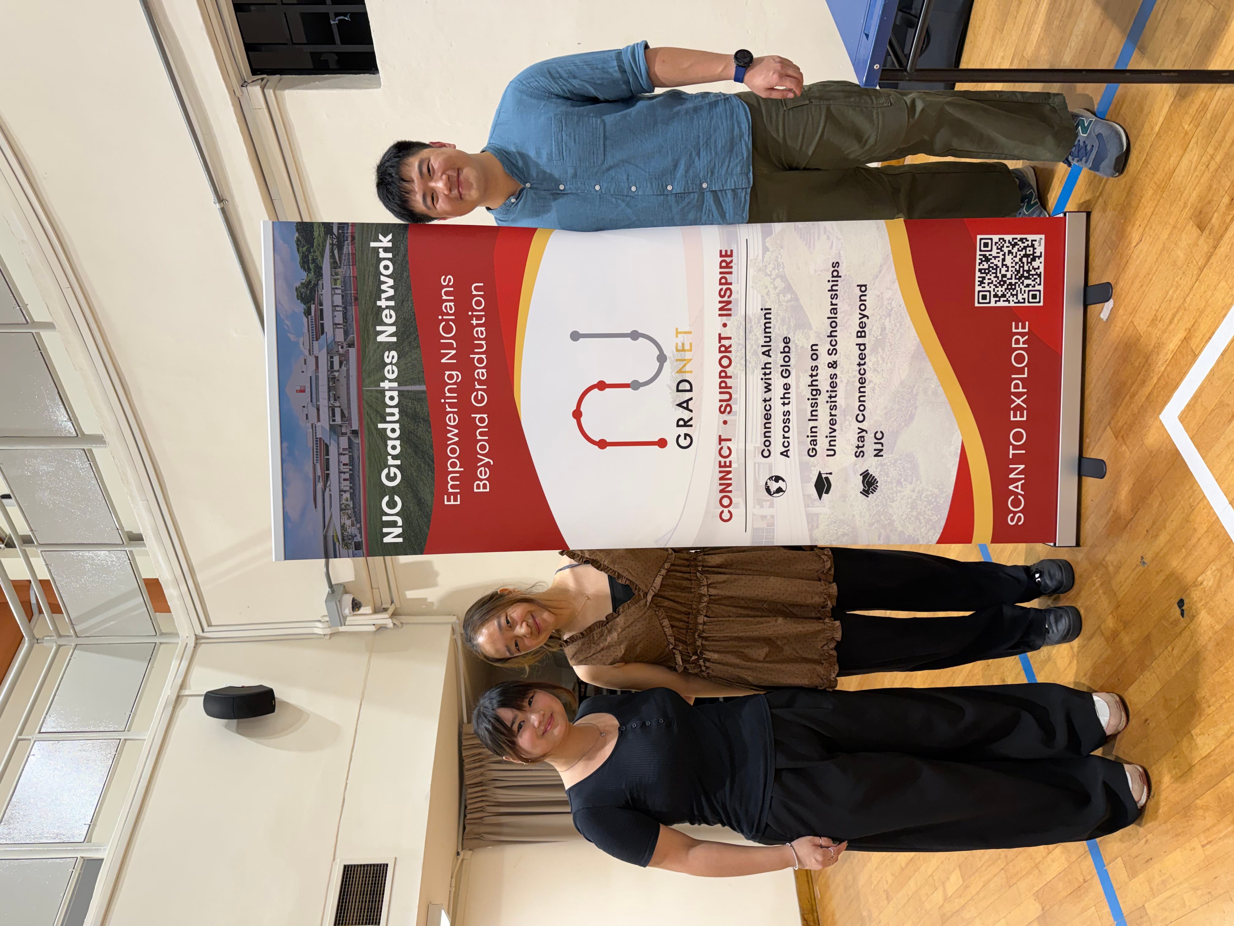 Three people stand beside an NJC Graduates Network GradNet banner in a hall with wooden floors.
