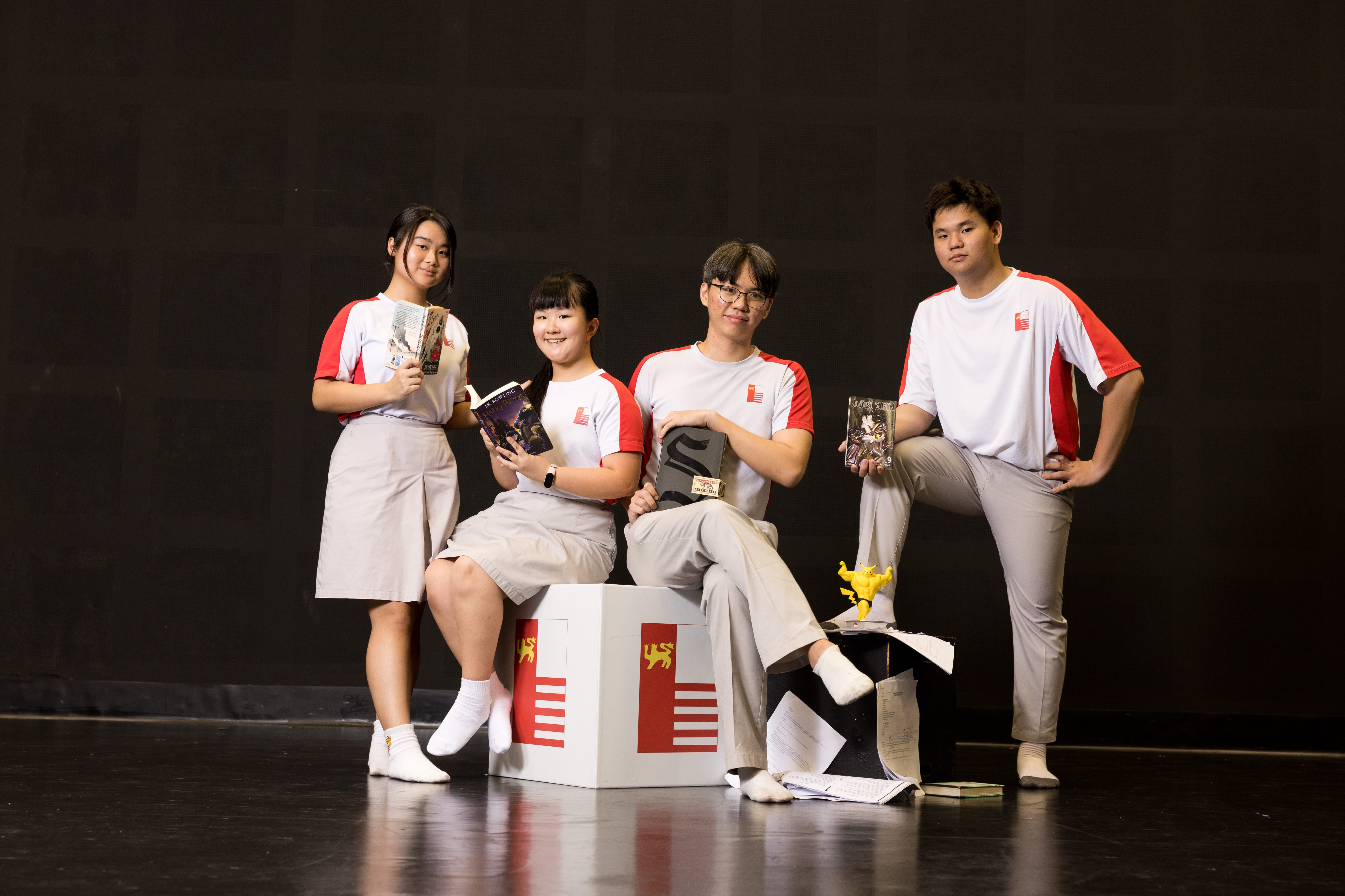 Four students in white-and-red uniforms pose with books on a stage against a black backdrop.