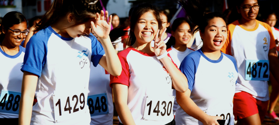 Young runners in numbered bibs smile and flash peace signs during a cheerful outdoor race event.