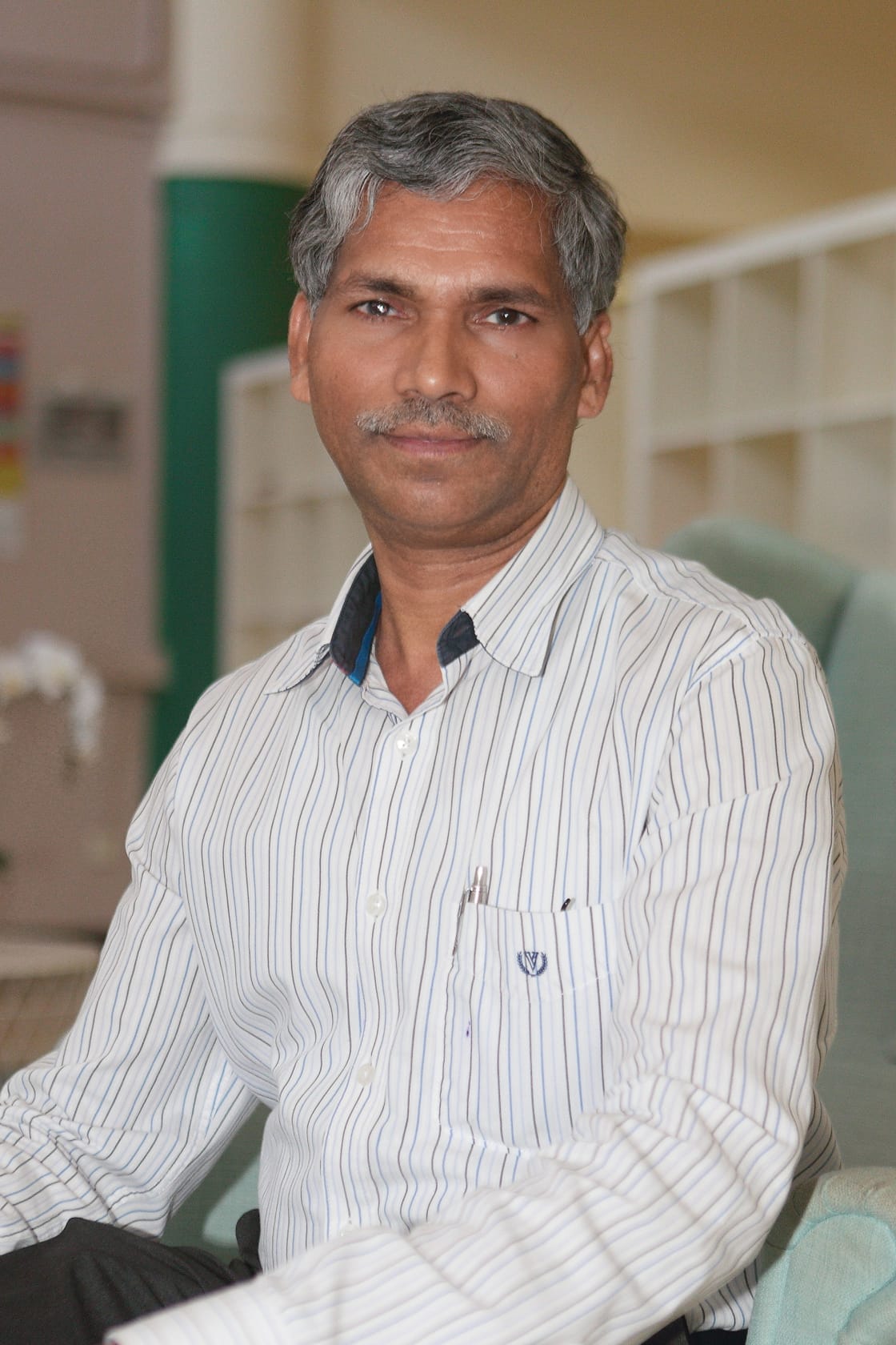 Middle-aged man with gray hair and mustache wearing a white striped shirt, seated indoors, looking at camera.
