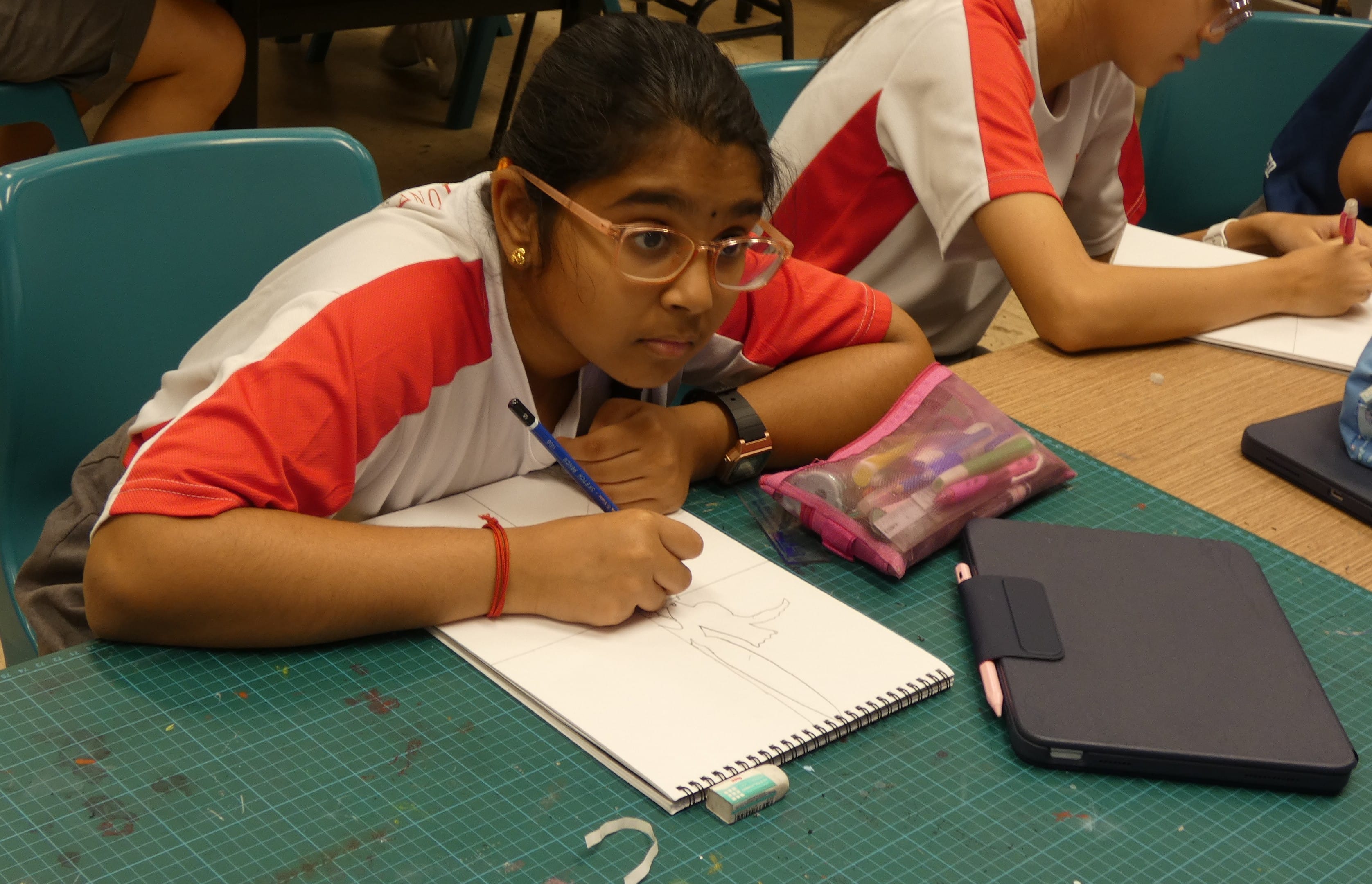 A young girl with glasses draws in a sketchbook at a green cutting mat desk in a classroom.