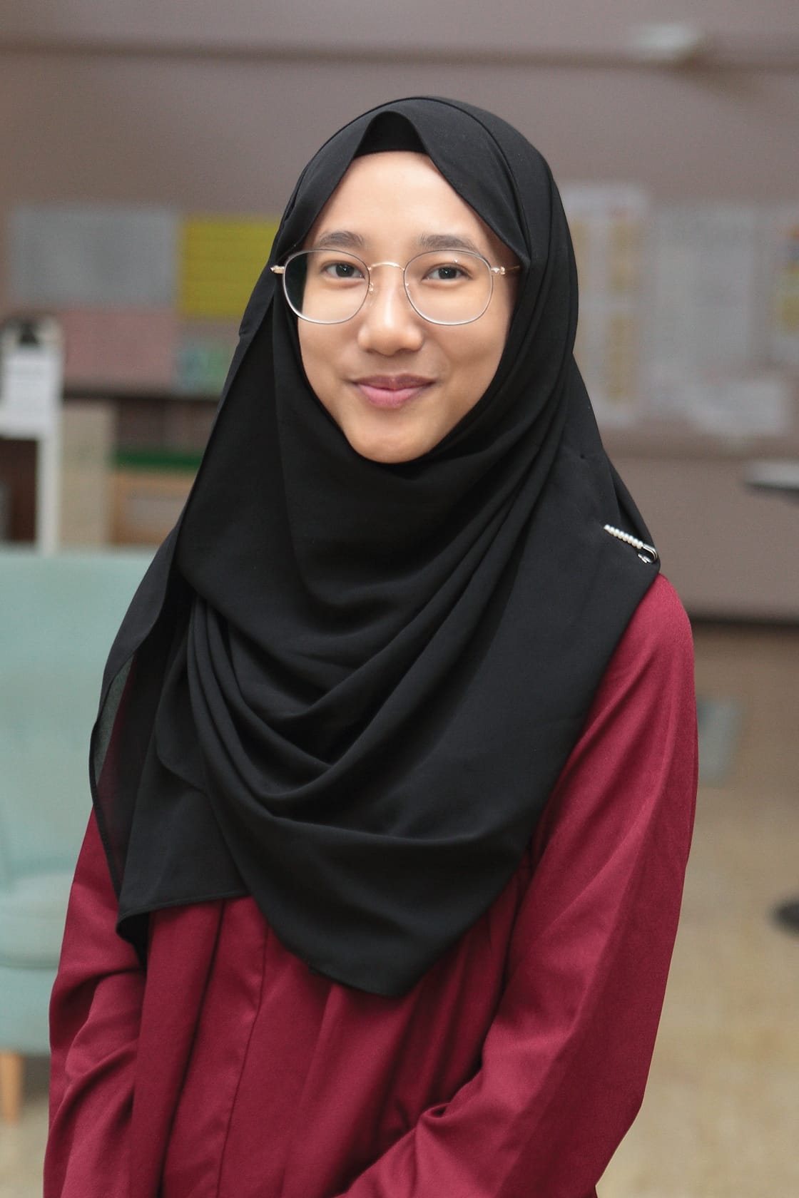 A young woman wearing a black hijab, round glasses, and a dark red top smiles in a blurred indoor setting.