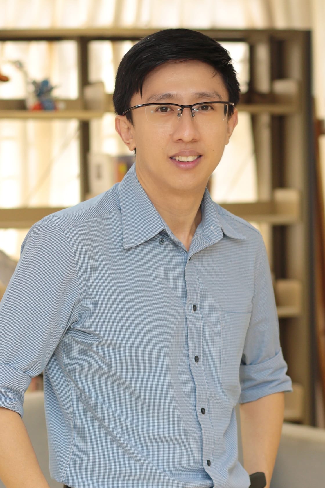 A smiling young man with glasses and short black hair wearing a light blue button-up shirt, standing in front of a bookshelf.