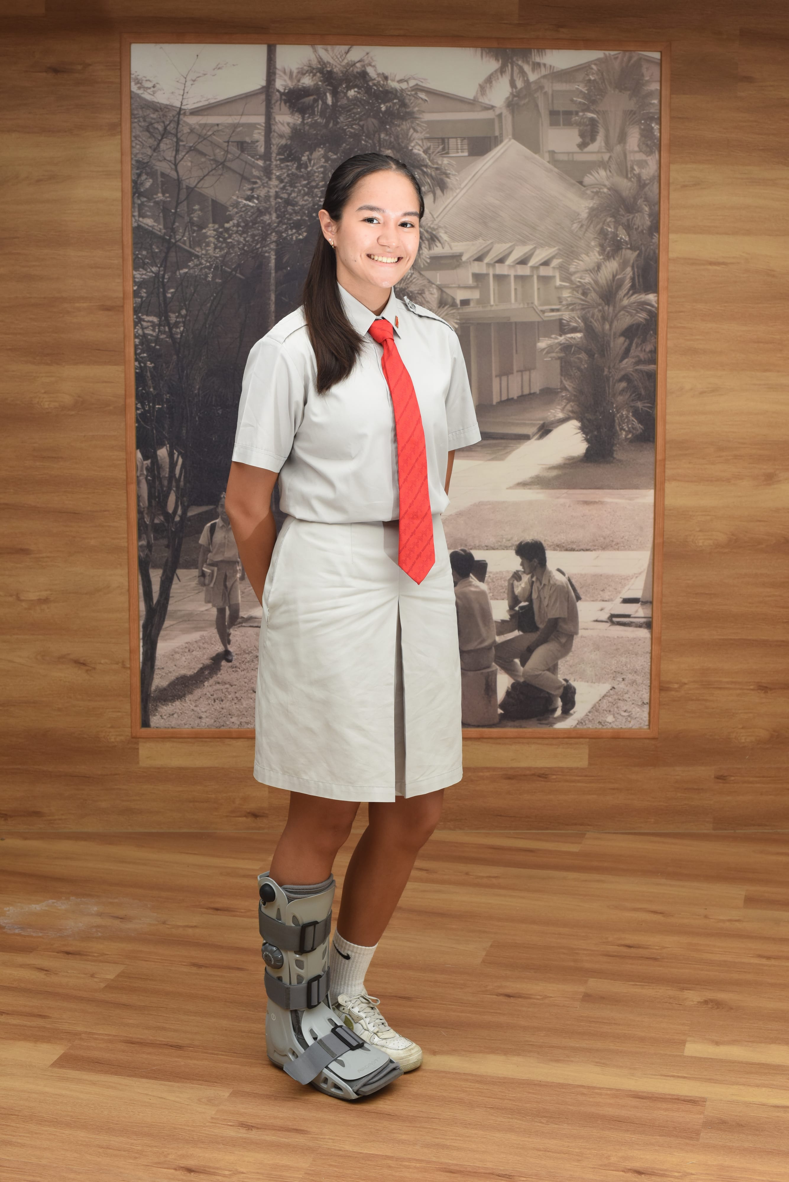 Smiling girl in white school uniform with red tie, wearing a medical boot on one leg, standing before a black-and-white photo.