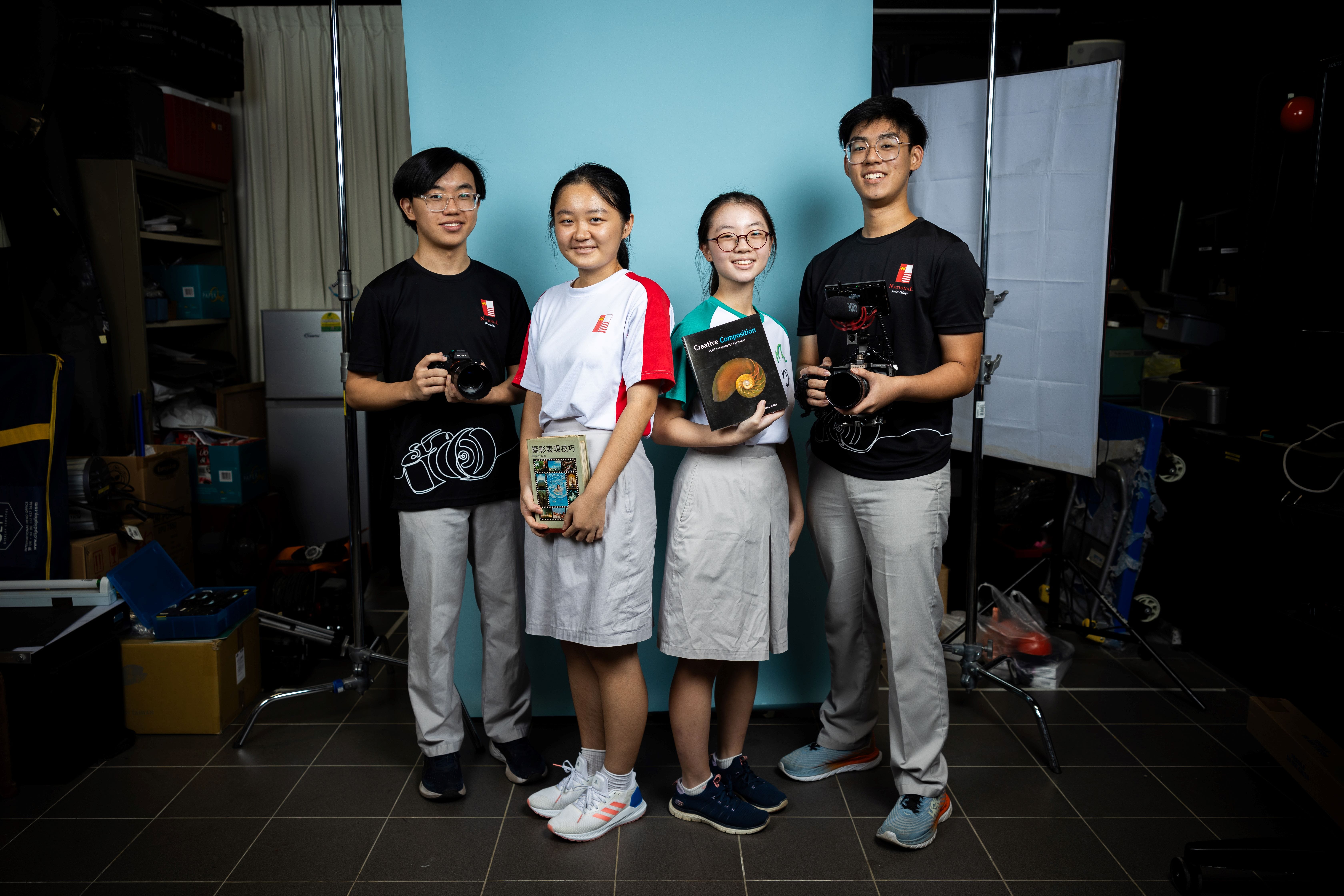 Four young students smiling, two holding cameras and two holding photography books, standing before a teal backdrop.