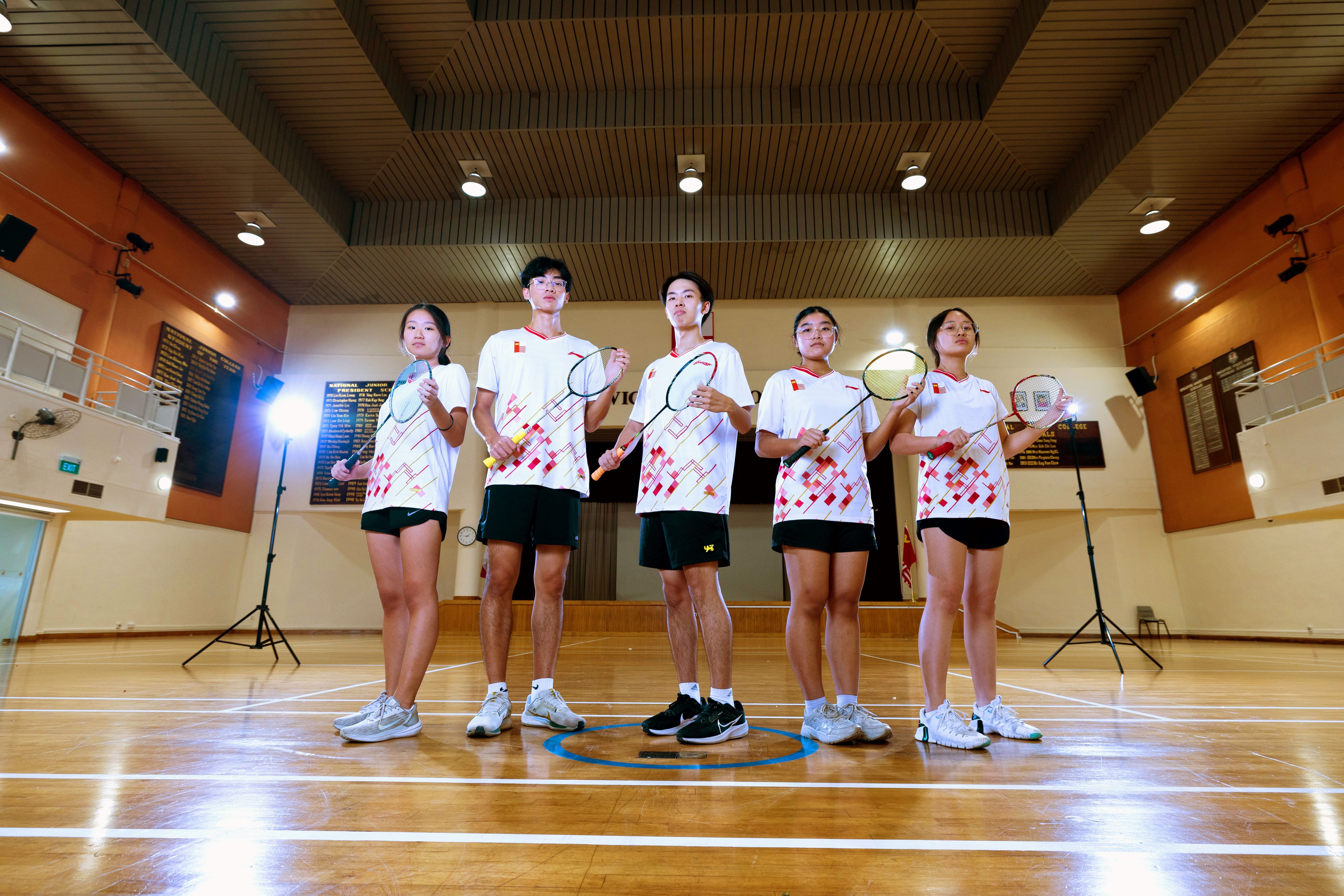 Five badminton players in matching white jerseys and black shorts pose with rackets in a gymnasium.