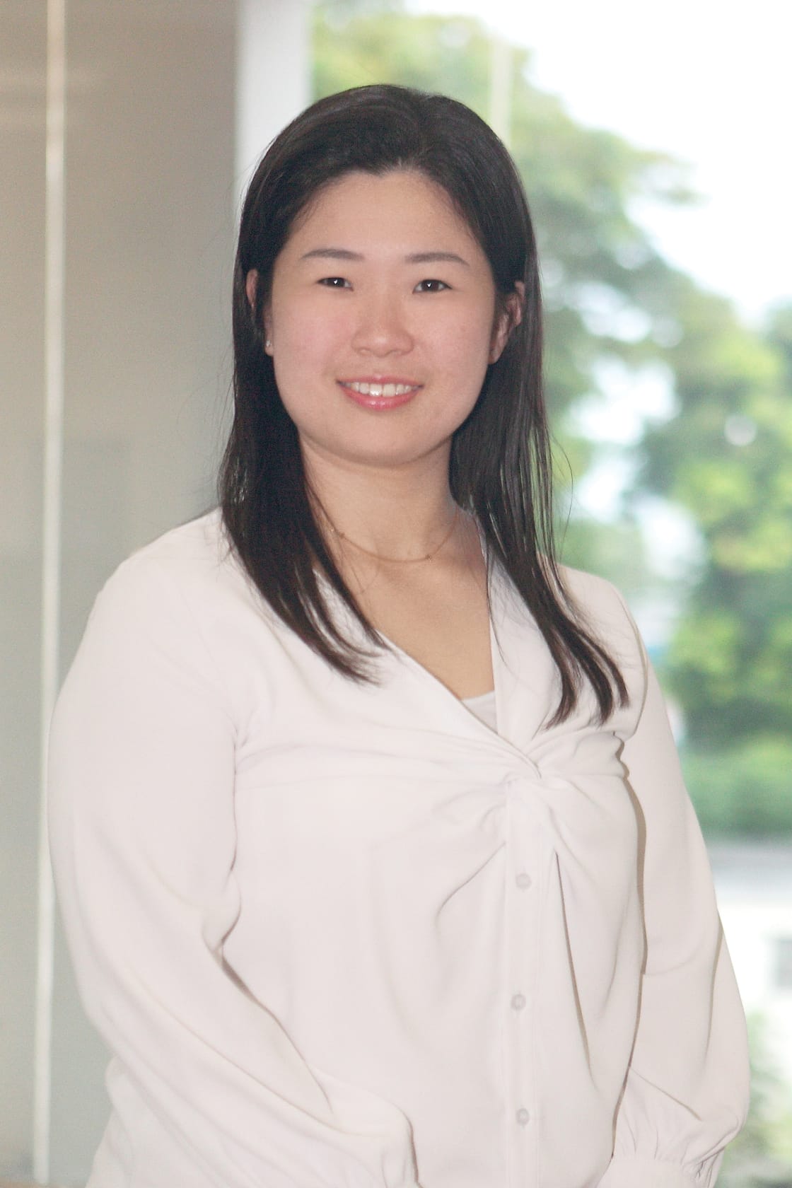 A smiling woman with long dark hair wearing a white blouse, standing indoors with blurred green trees in the background.