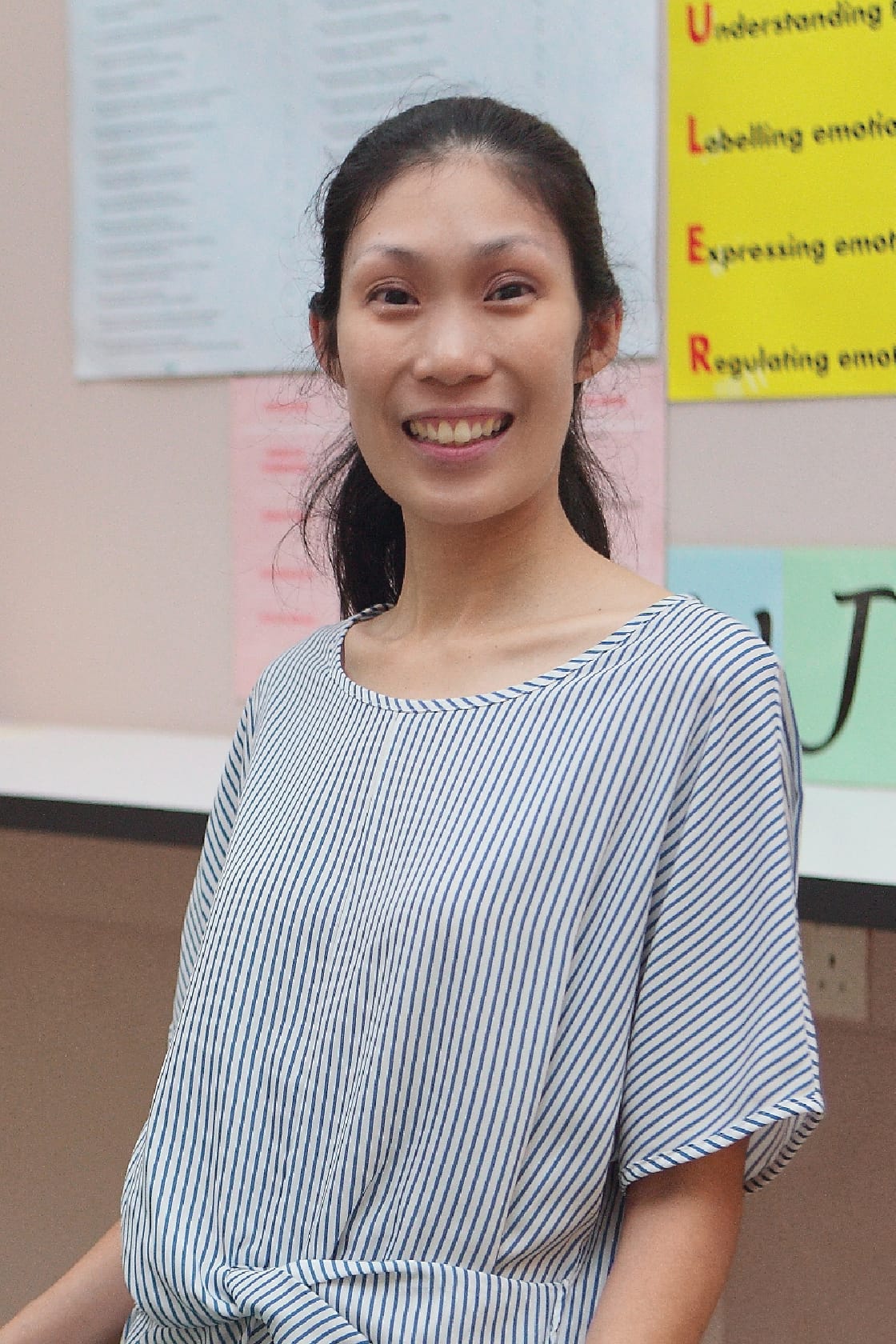 A smiling young woman with dark hair in a striped top stands before colorful posters about emotions on a wall.