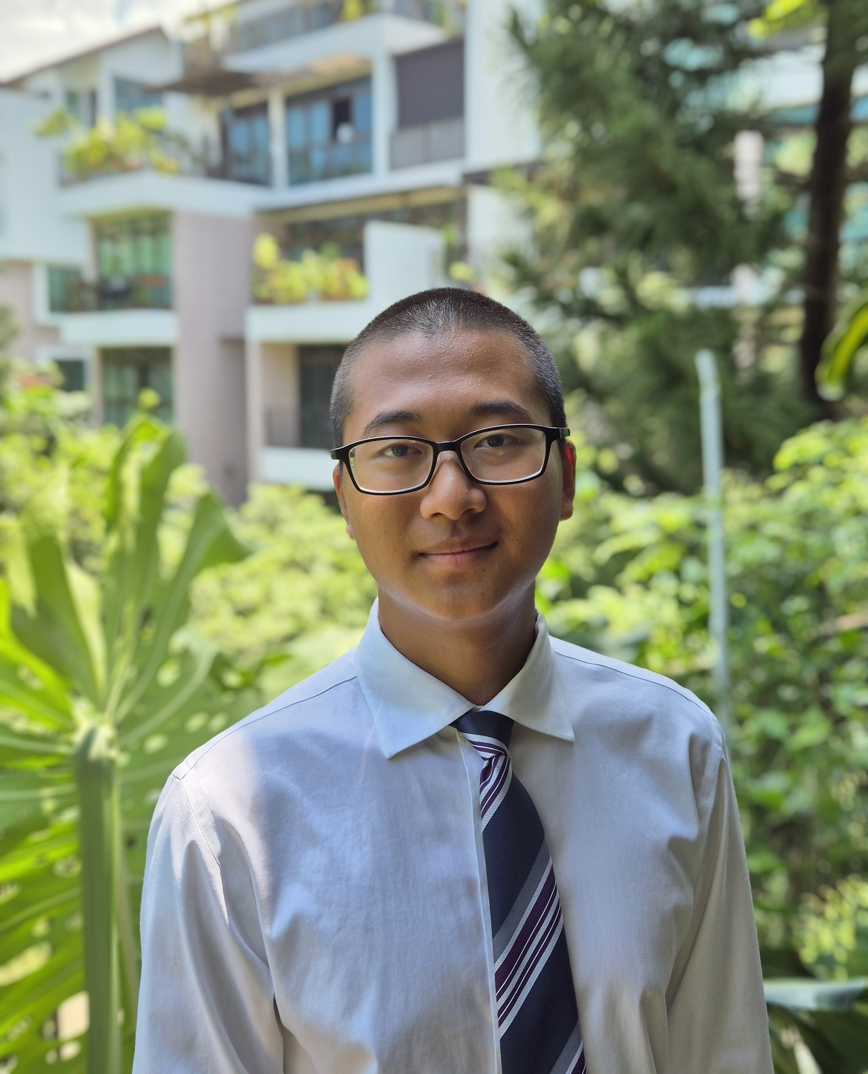 Young man with glasses, short hair, white dress shirt and striped tie, standing outdoors with green trees and a building behind him.